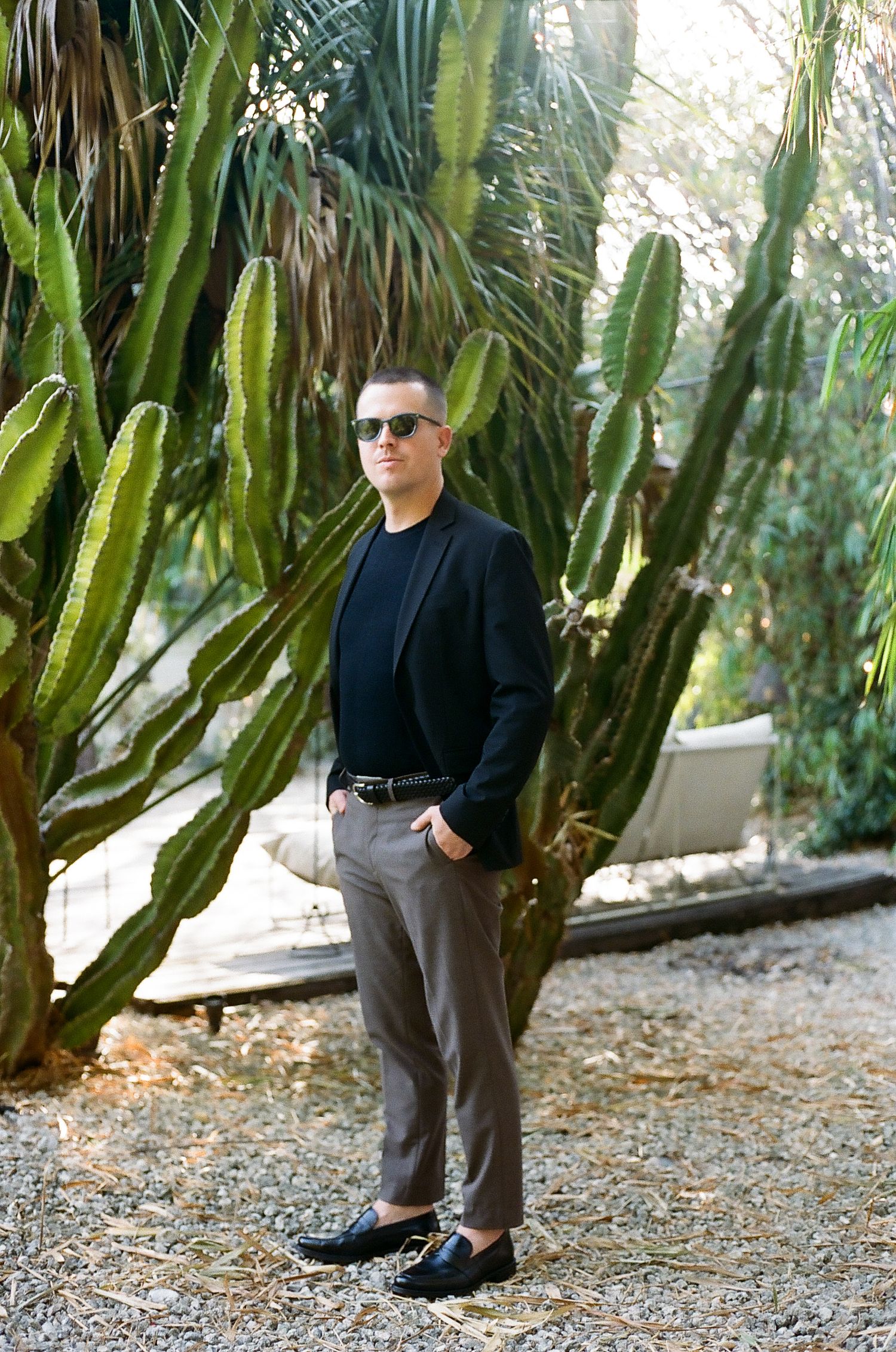 Figure in black clothing standing among tall cacti and tropical plants in a garden setting.