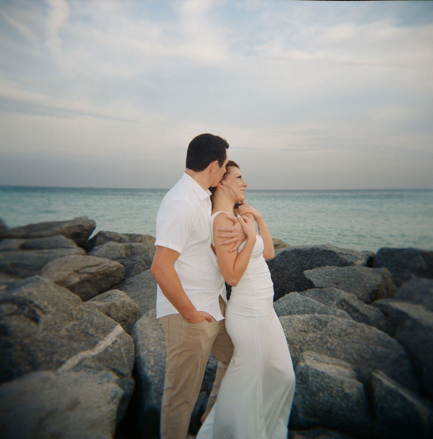 A romantic couple embraces on rocky coastal outcrop against turquoise waters at sunset.