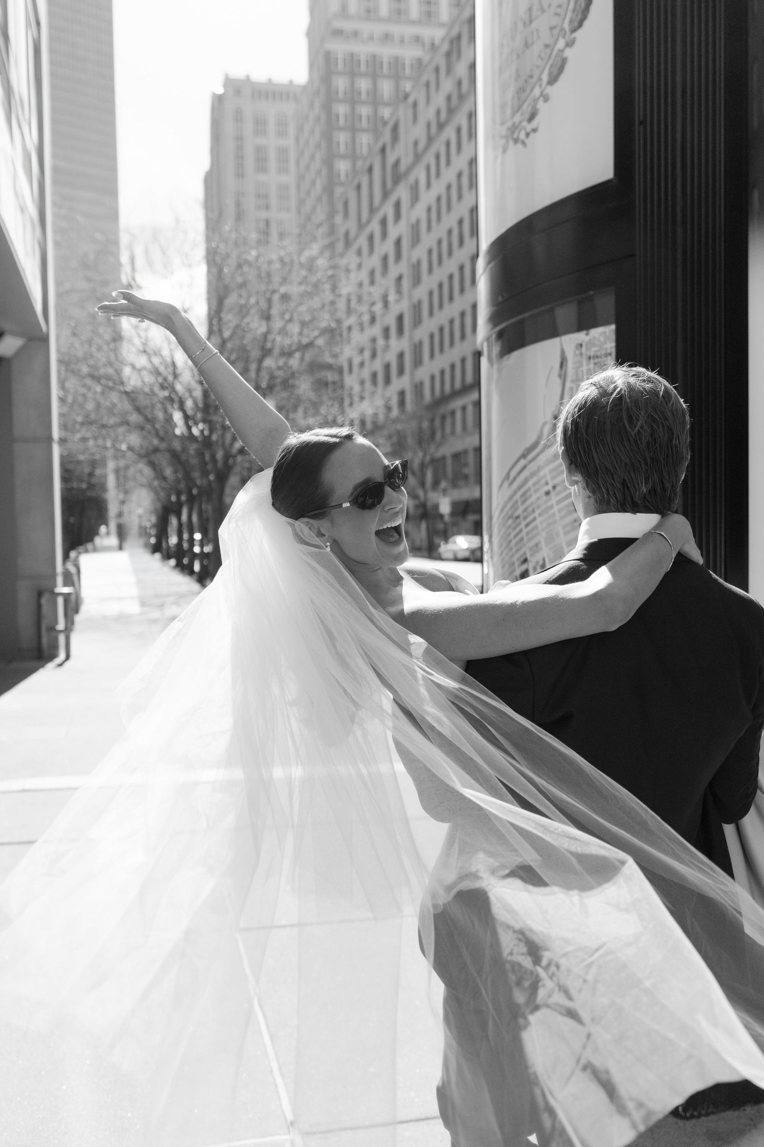 Joyful bride in sunglasses and flowing veil raises her arm while groom holds her on a sunny city sidewalk.
