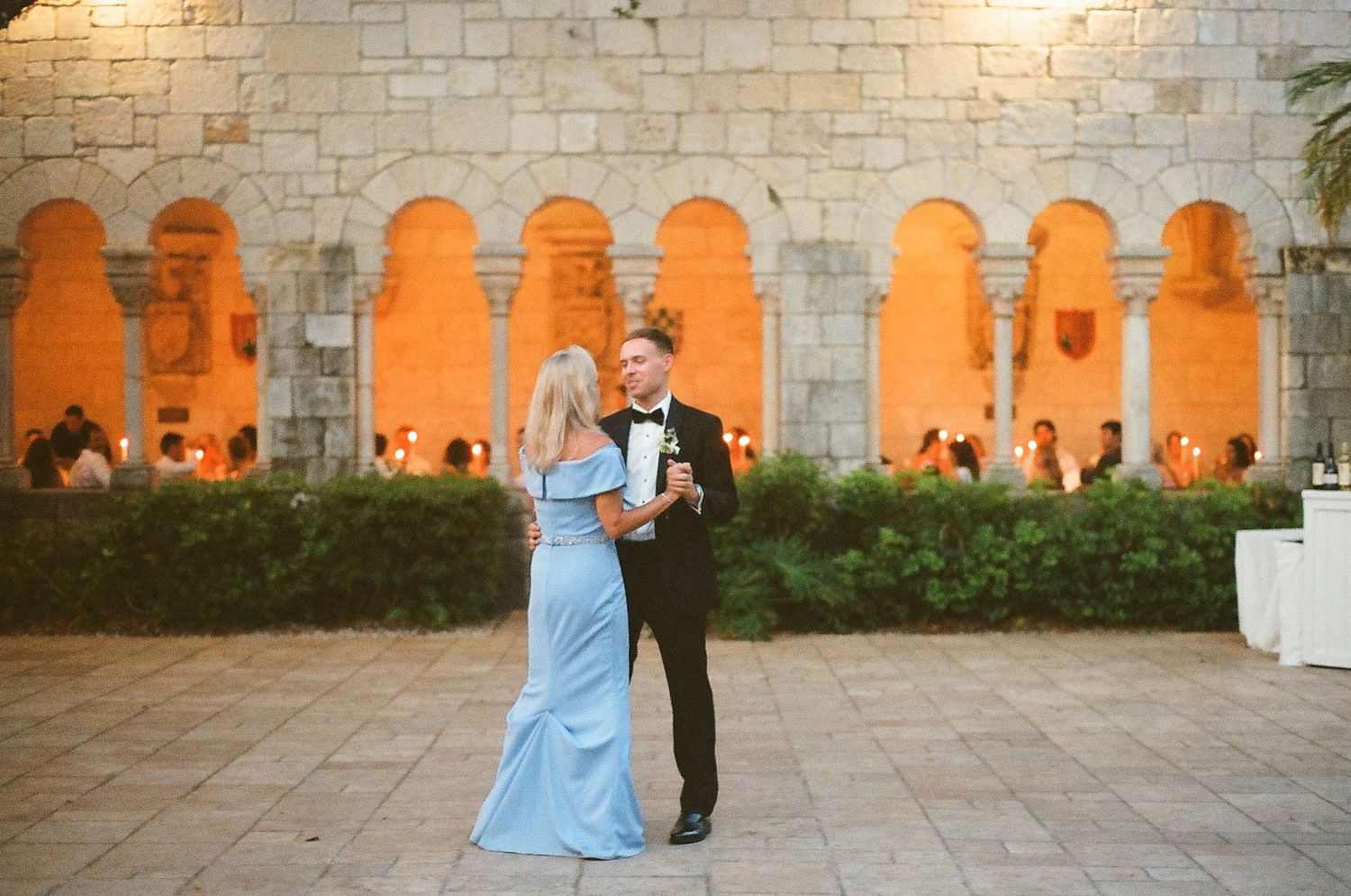 Couple dancing in elegant stone courtyard with glowing arches and wedding guests in background at night.