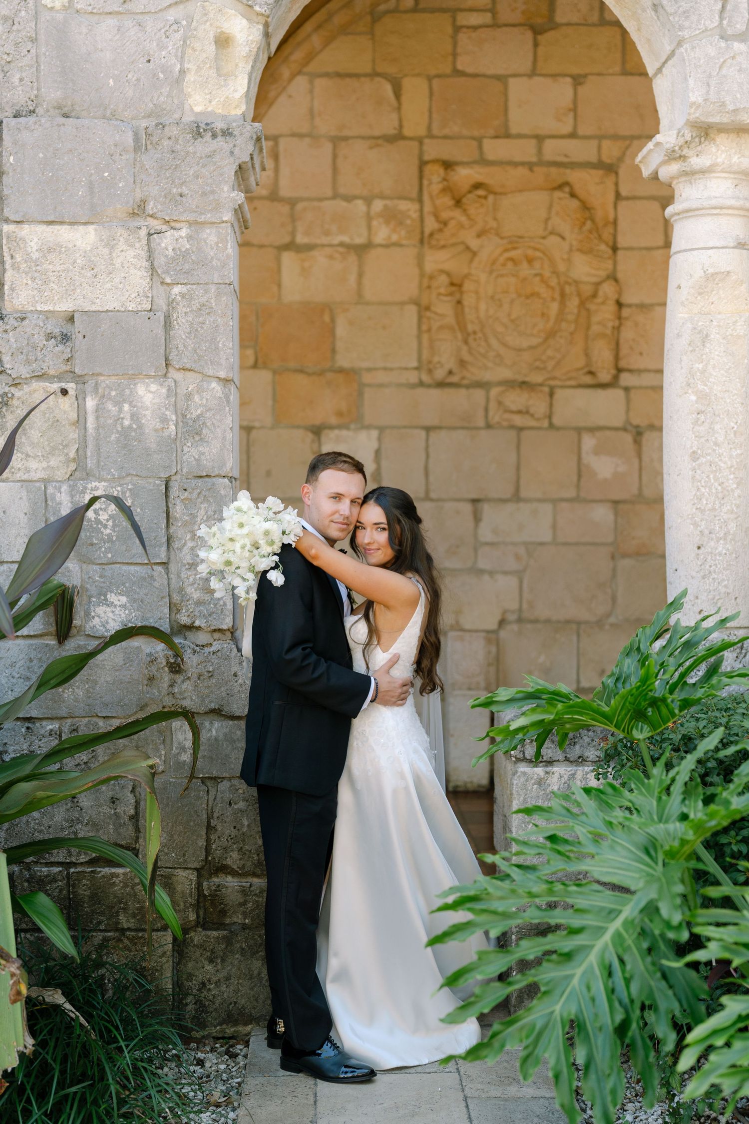 Bride and groom embracing before a stone arch with carved crest, surrounded by tropical greenery.