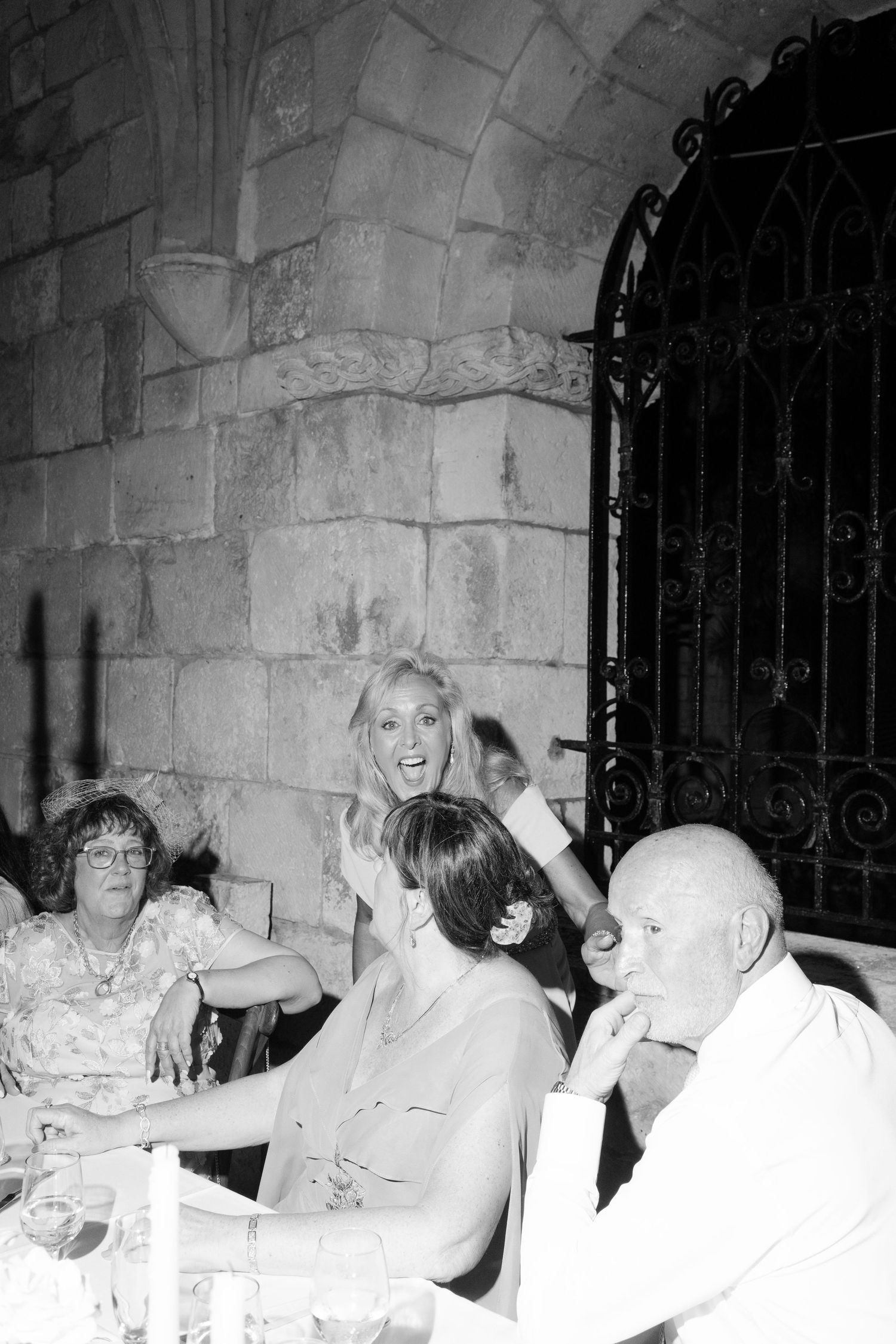 Black and white candid photo of wedding guests laughing and socializing at reception table beside stone wall.