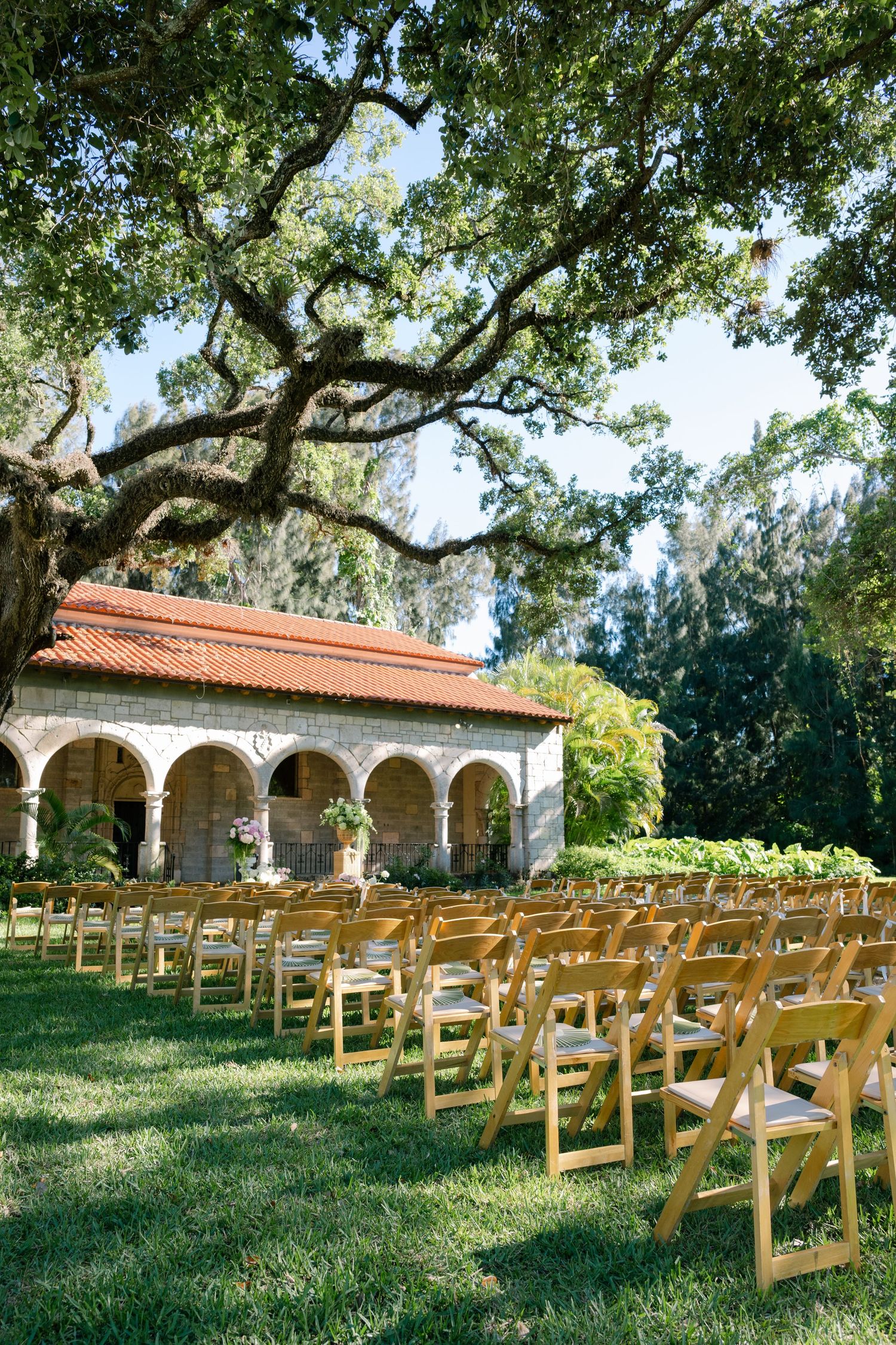 Outdoor wedding ceremony setup with wooden chairs on green lawn beneath oak tree with Spanish-style building backdrop.