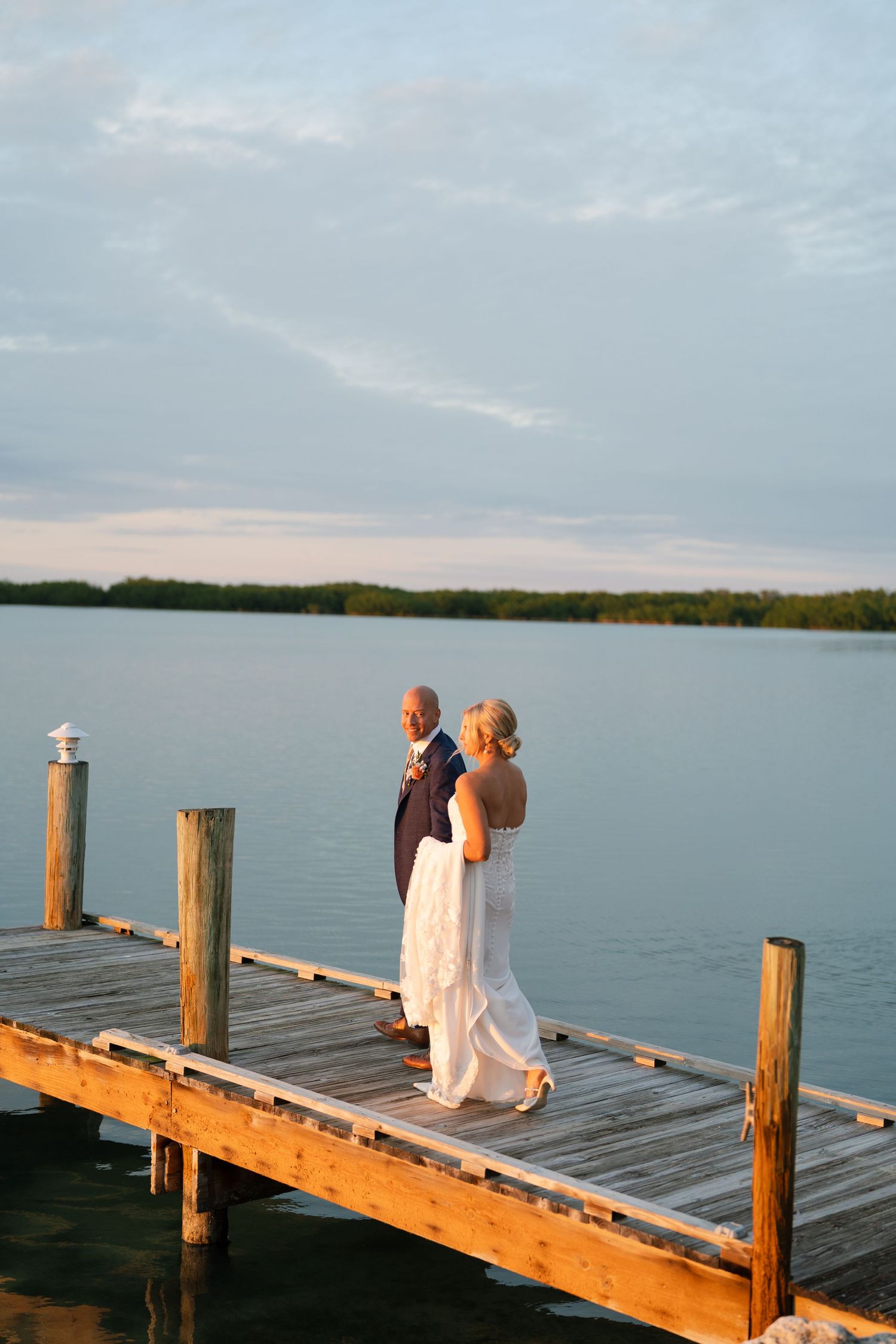 Two figures standing on a wooden dock at sunset overlooking a serene lake.