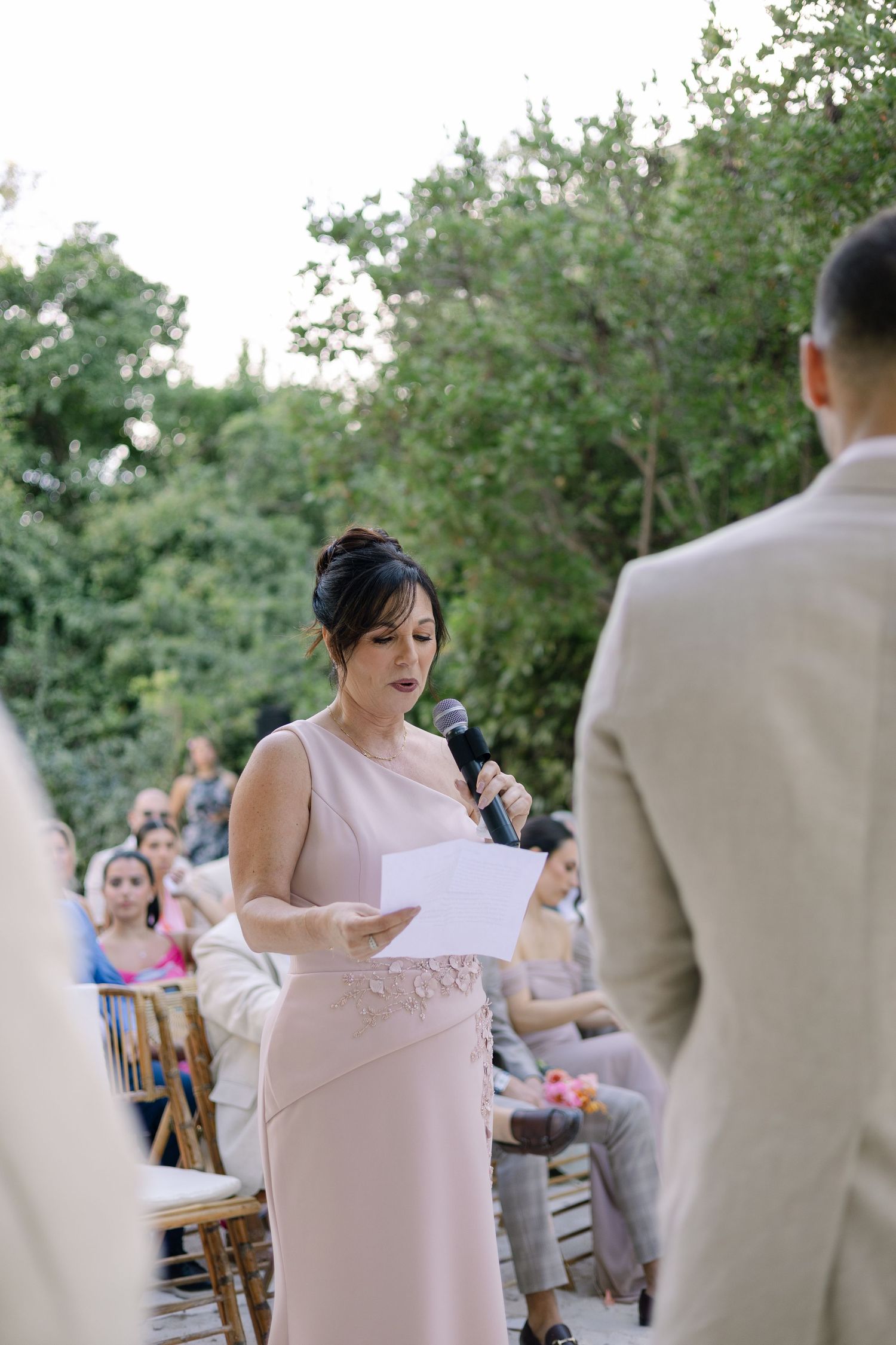 An outdoor wedding ceremony with someone in pink reading from papers while guests watch from wooden chairs.