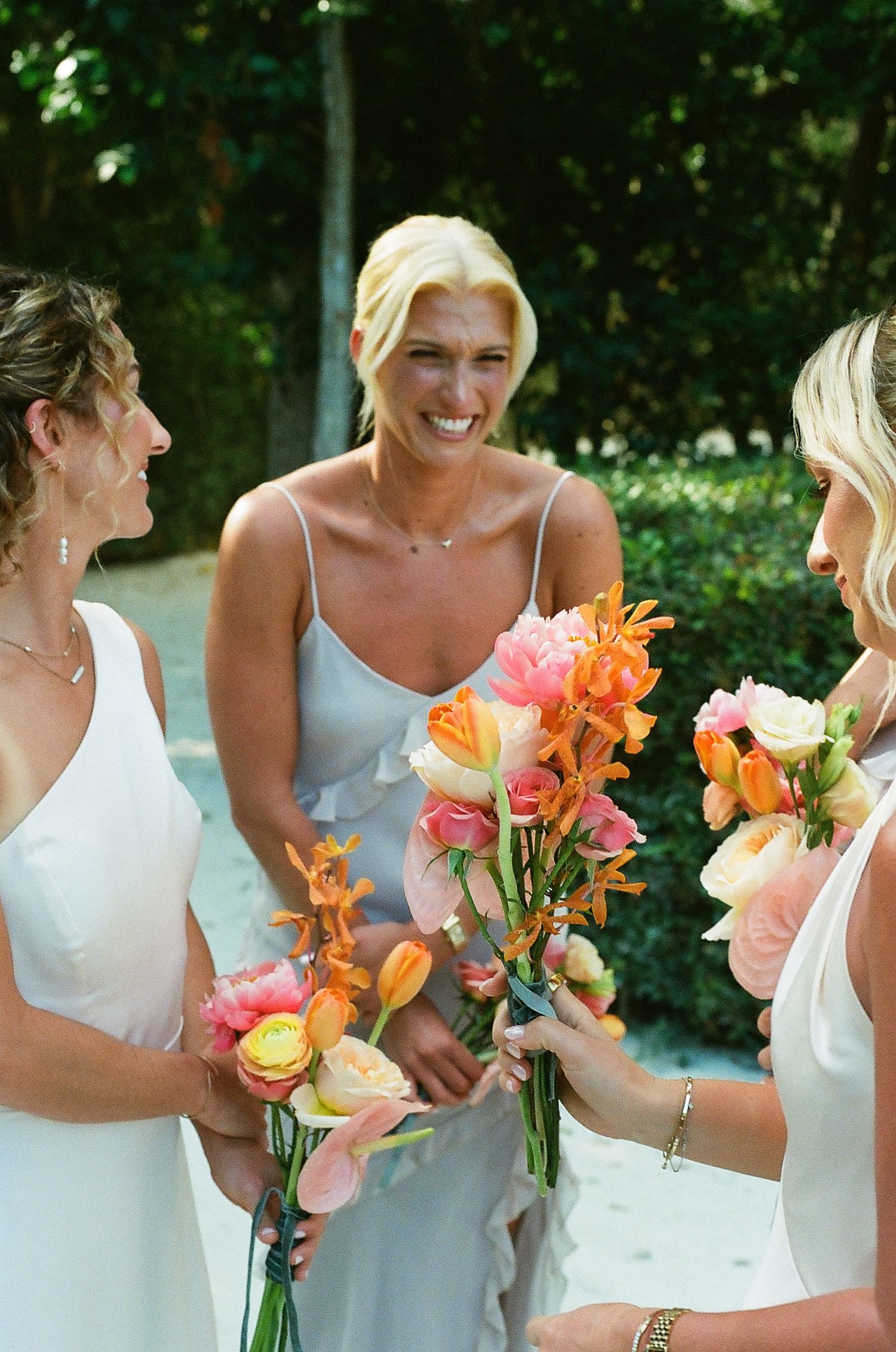 Bridesmaids in white dresses hold vibrant orange and pink floral bouquets during an outdoor wedding celebration.