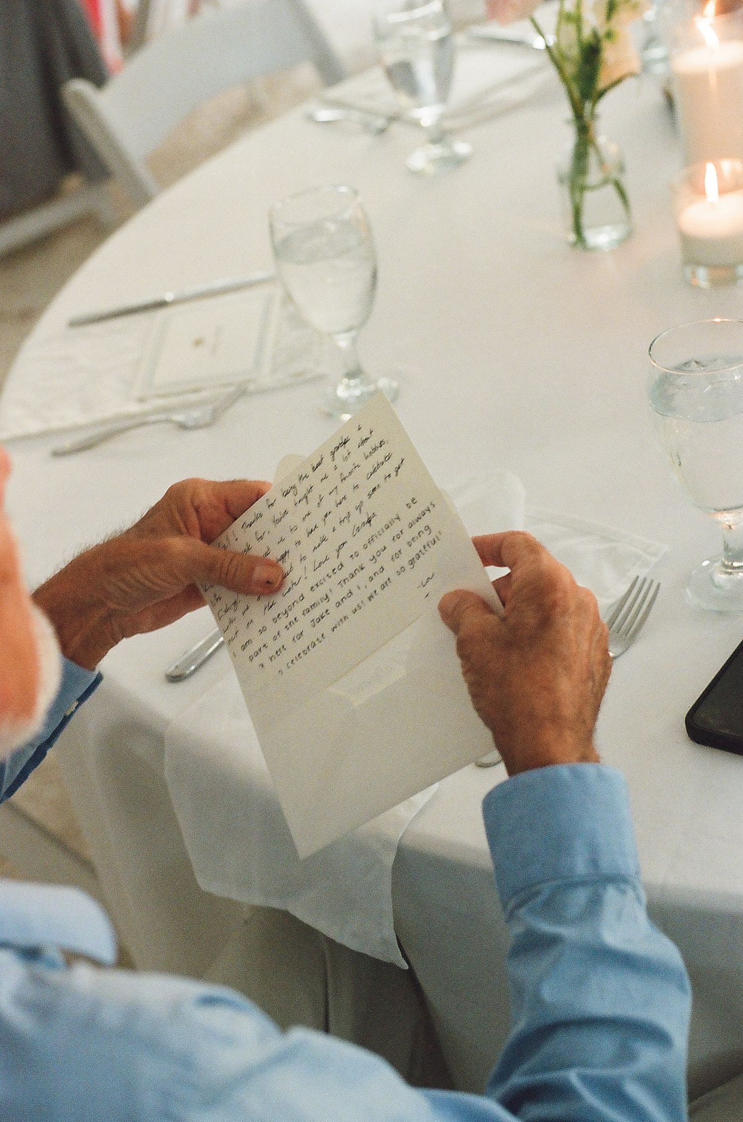 Elderly man in blue shirt reading handwritten letter at elegant wedding reception table with candles and glassware.