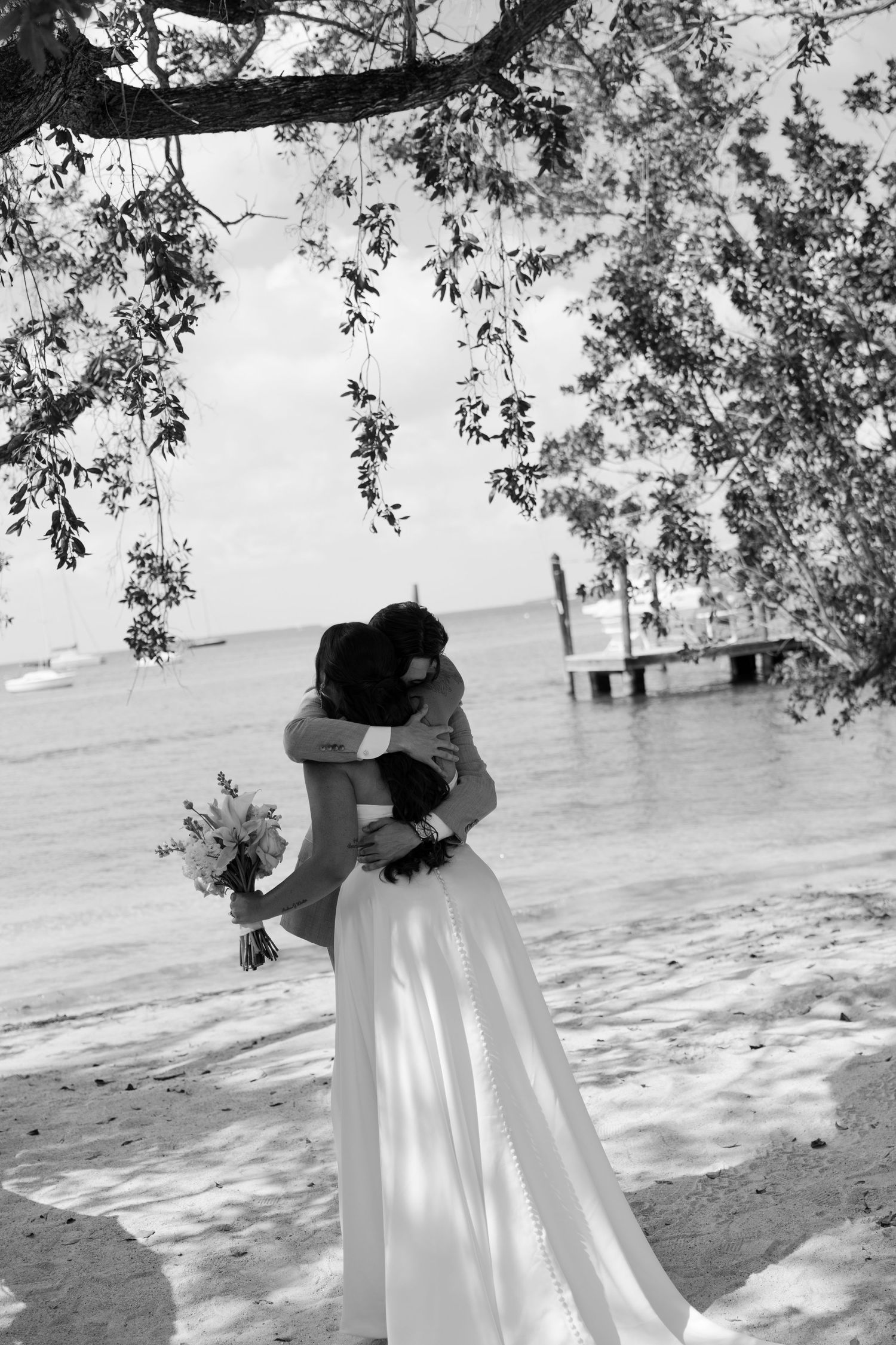 A black and white image of a couple embracing on a beach with trees in the background.
