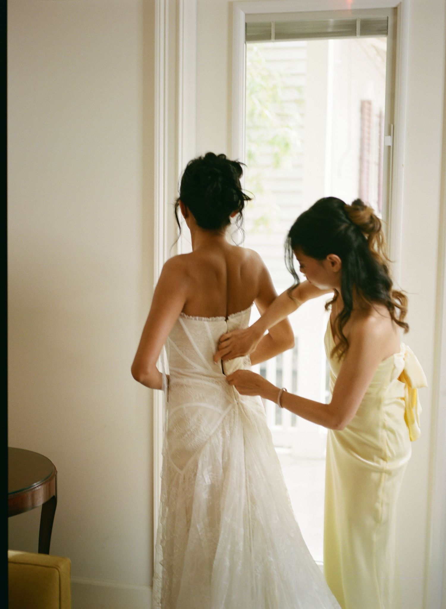 Bridesmaid in yellow dress helping bride button up white wedding gown near a bright window.