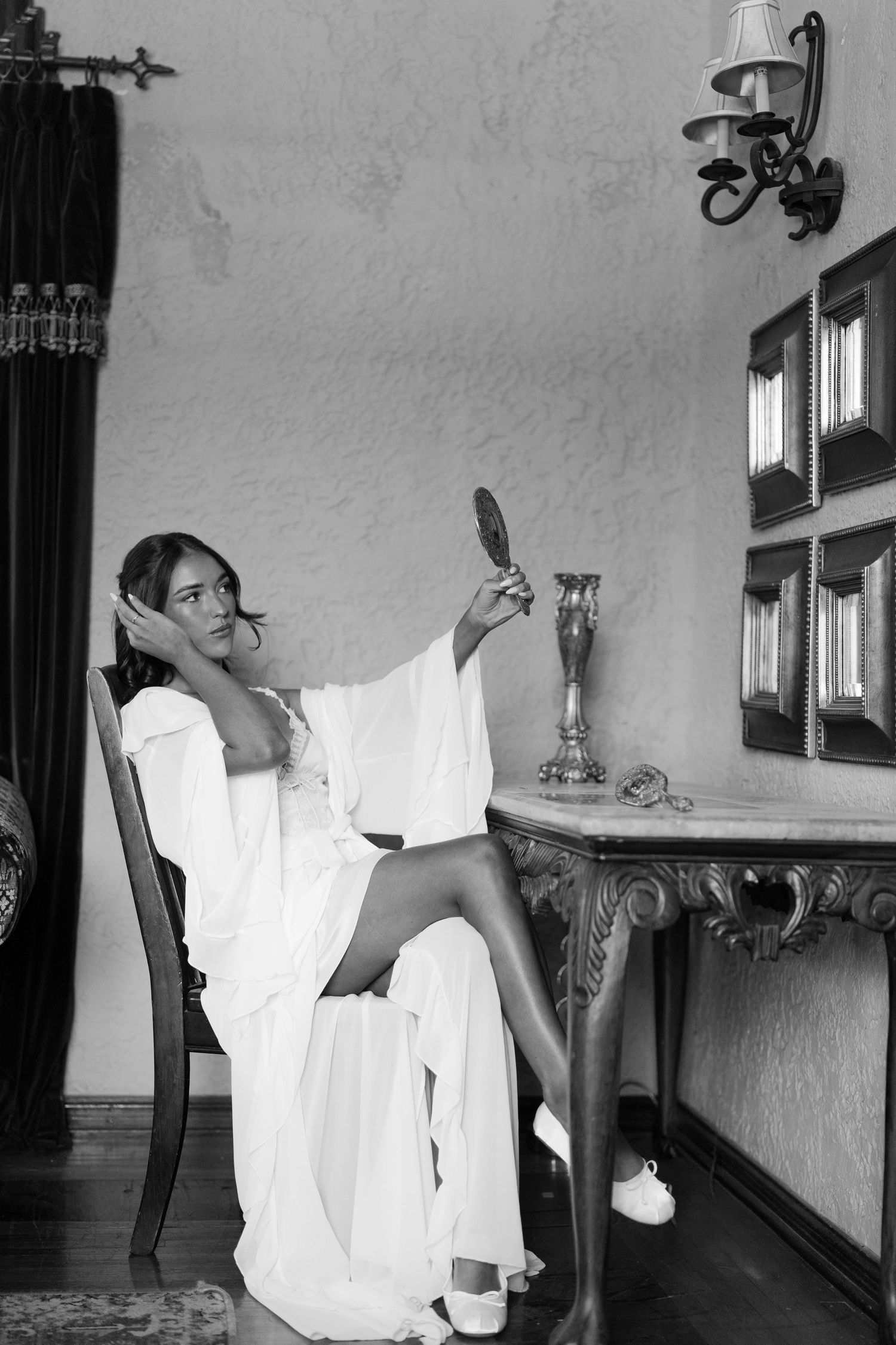 Elegant woman in white robe sits at vintage vanity table holding hand mirror in black and white photo.