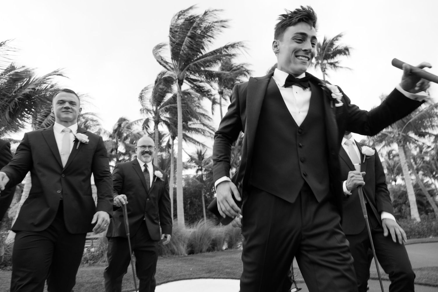 Groom and groomsmen in black tuxedos celebrate outdoors near palm trees on wedding day.