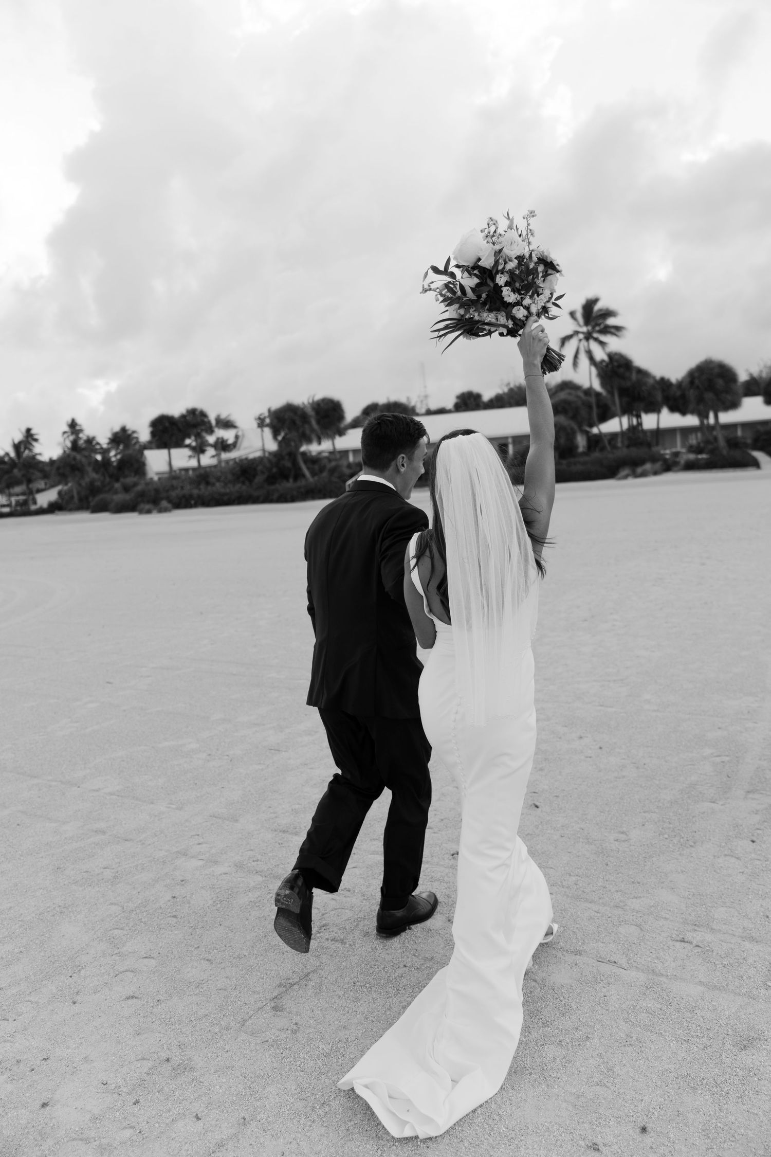 Bride in white gown raising bouquet with groom on sandy beach, black and white wedding photo.