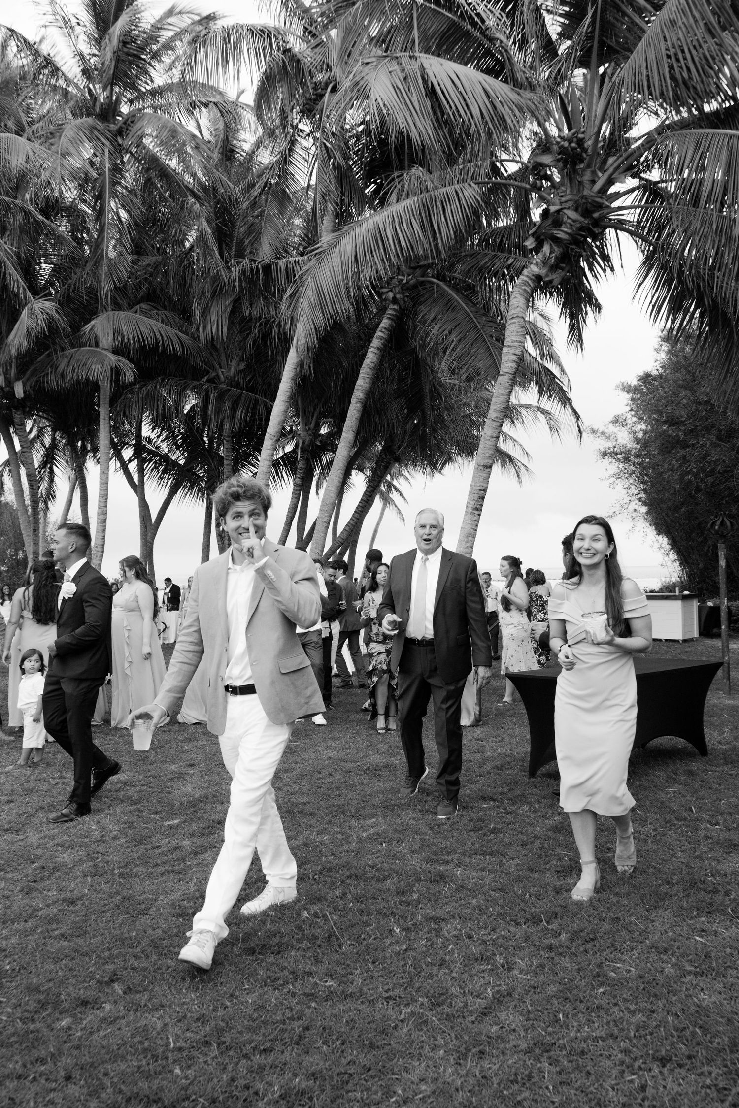 Black and white photo of wedding guests walking under palm trees at outdoor tropical reception.