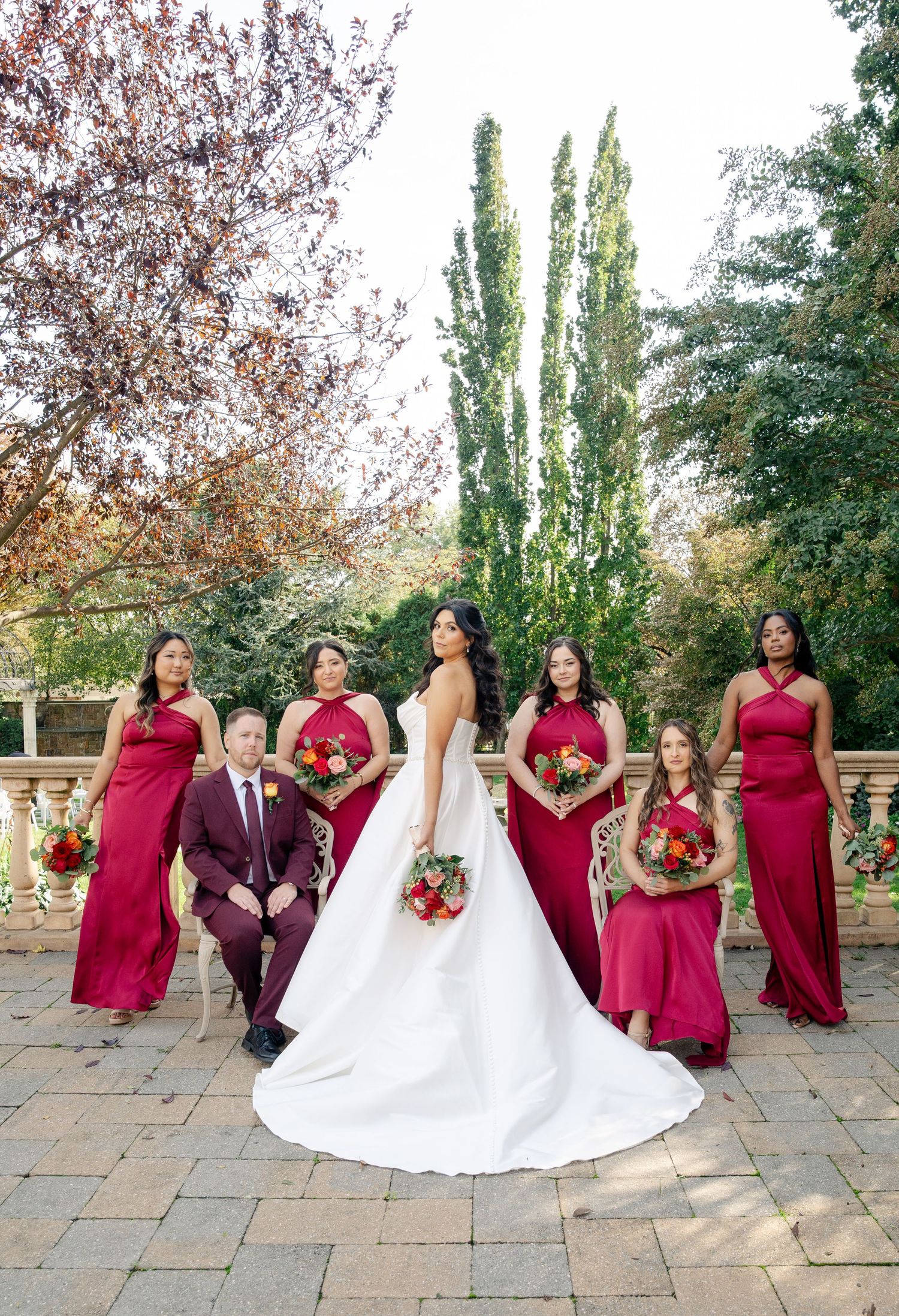 Wedding party in burgundy dresses and suits poses together on stone patio during autumn outdoor photo session.