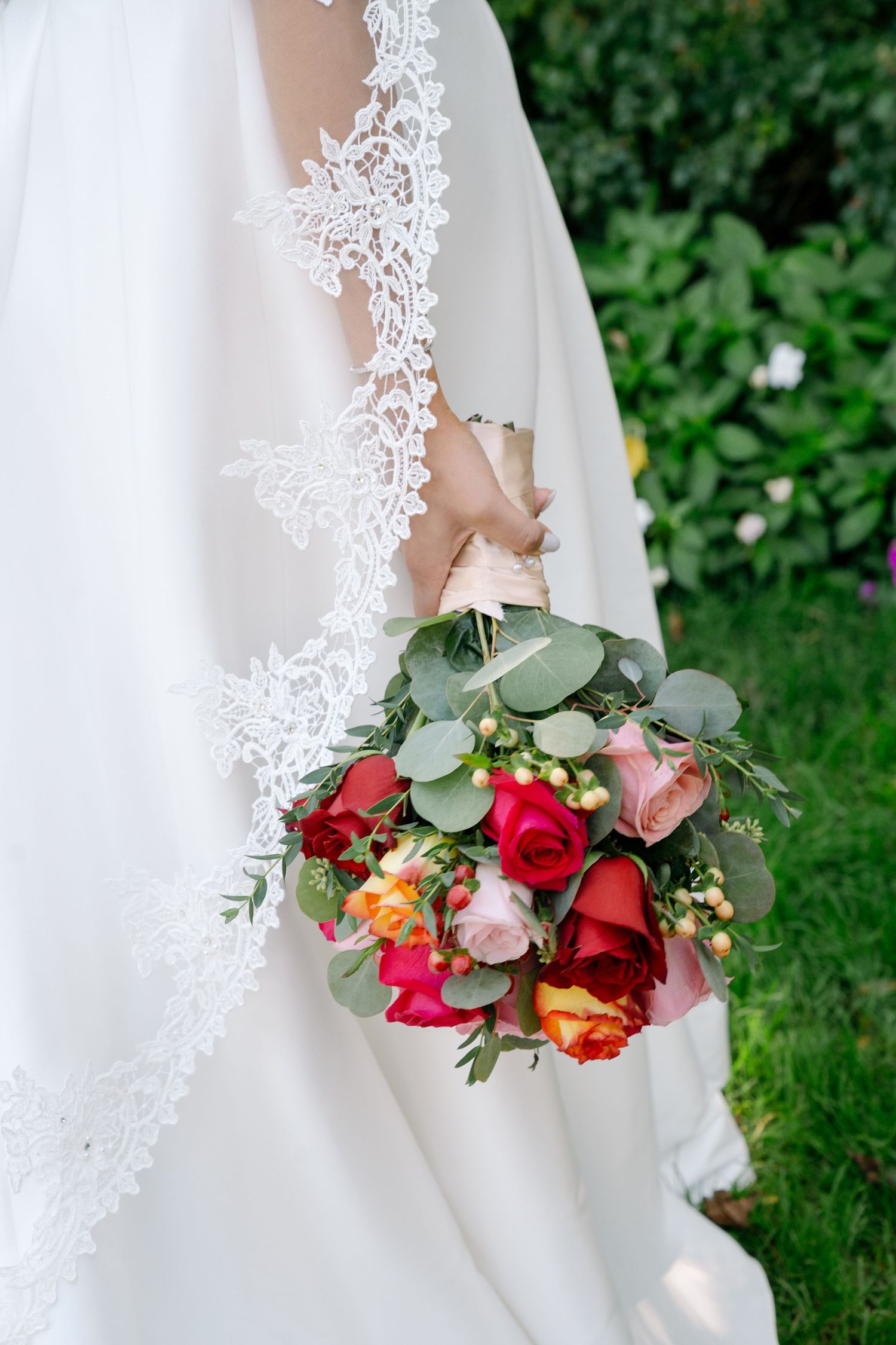 Delicate white lace wedding dress detail with vibrant red and coral floral bouquet against green foliage background.