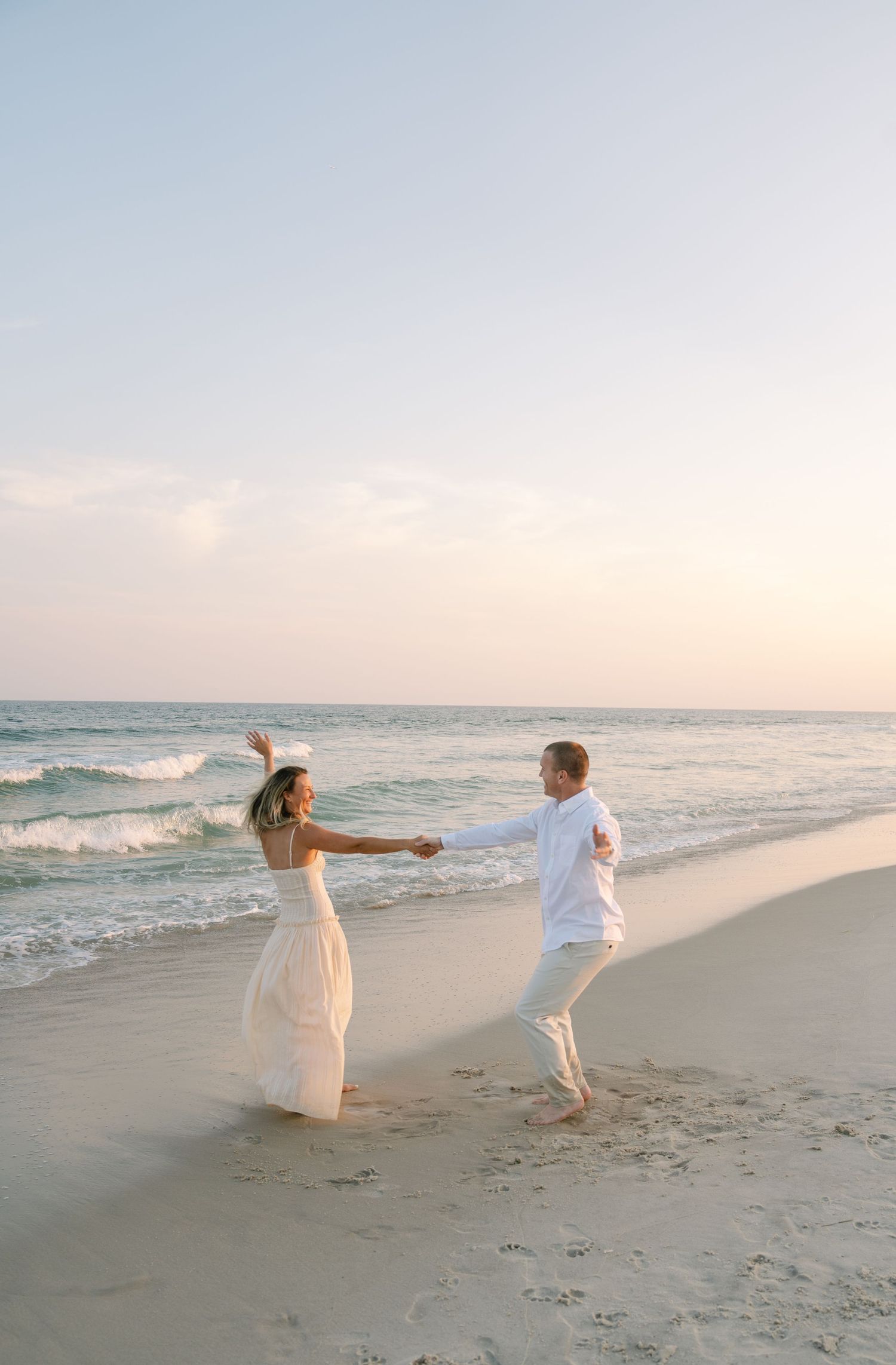 Two people in white outfits dance playfully together on a sandy beach at sunset with ocean waves in the background.