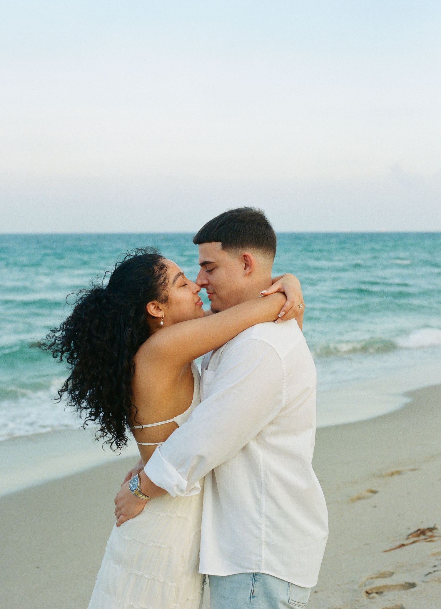 A couple shares a romantic moment on a scenic beach with turquoise waters and sandy shores in the background.