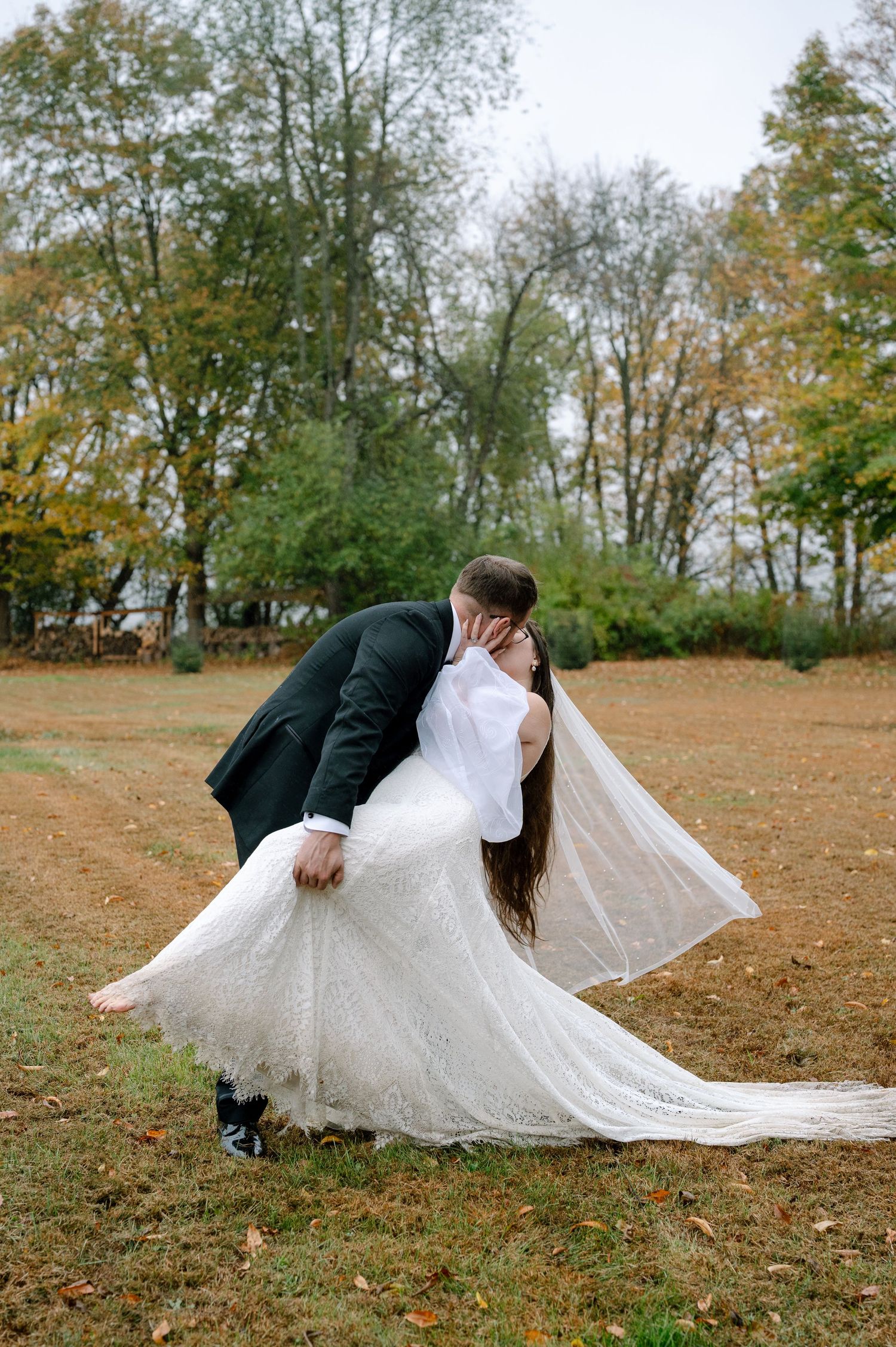 A couple in wedding attire lies dramatically on fallen autumn leaves in a park setting.