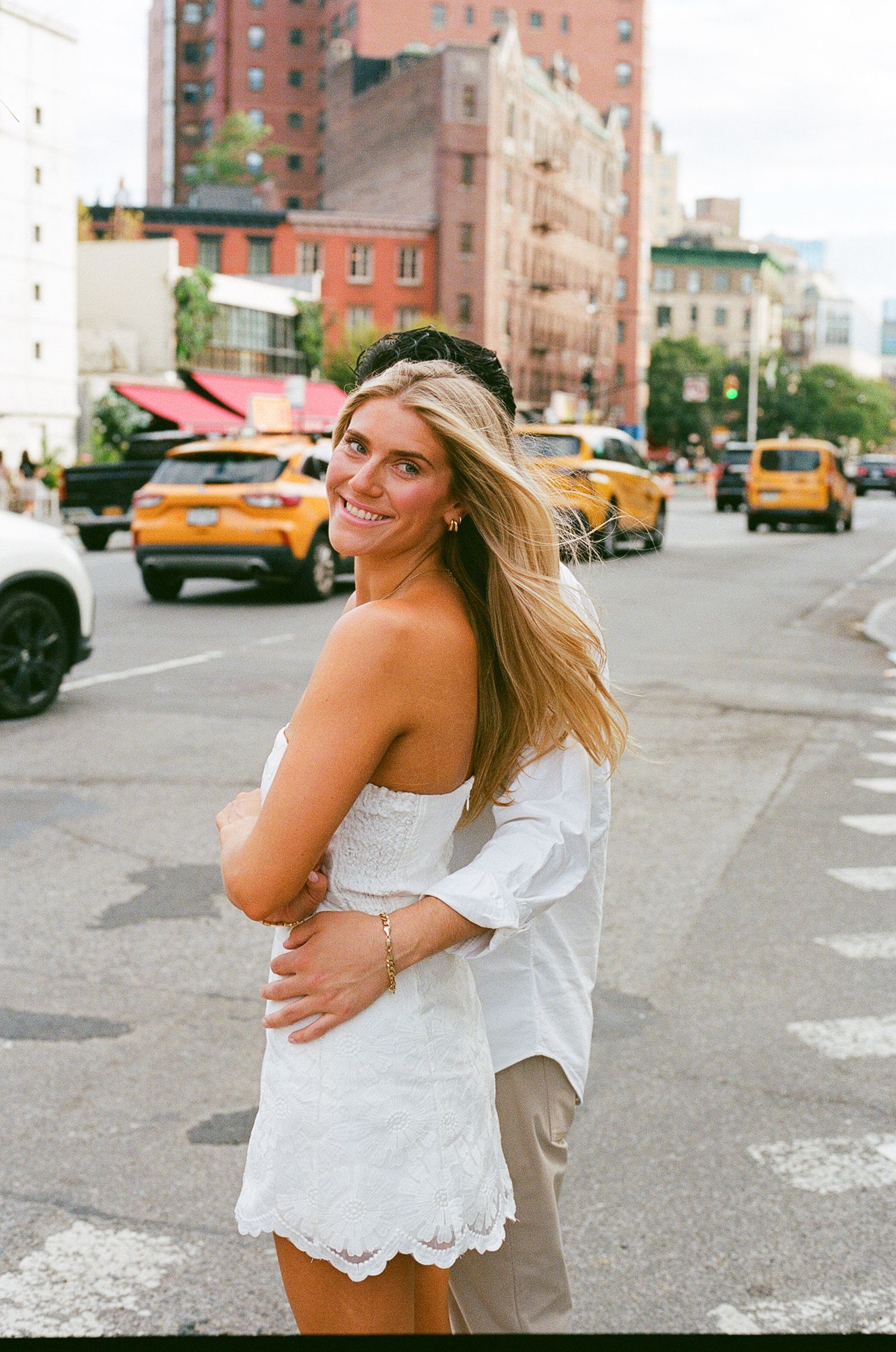 Smiling blonde woman in white lace outfit poses on a busy NYC street with yellow taxis in the background.
