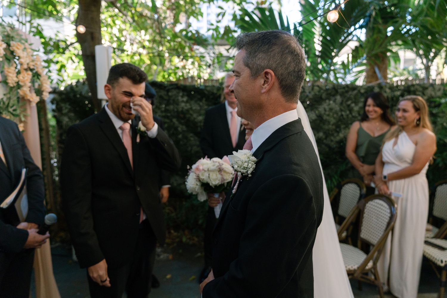 Groom wiping tears at outdoor wedding ceremony while man holds bridal bouquet near floral arch.