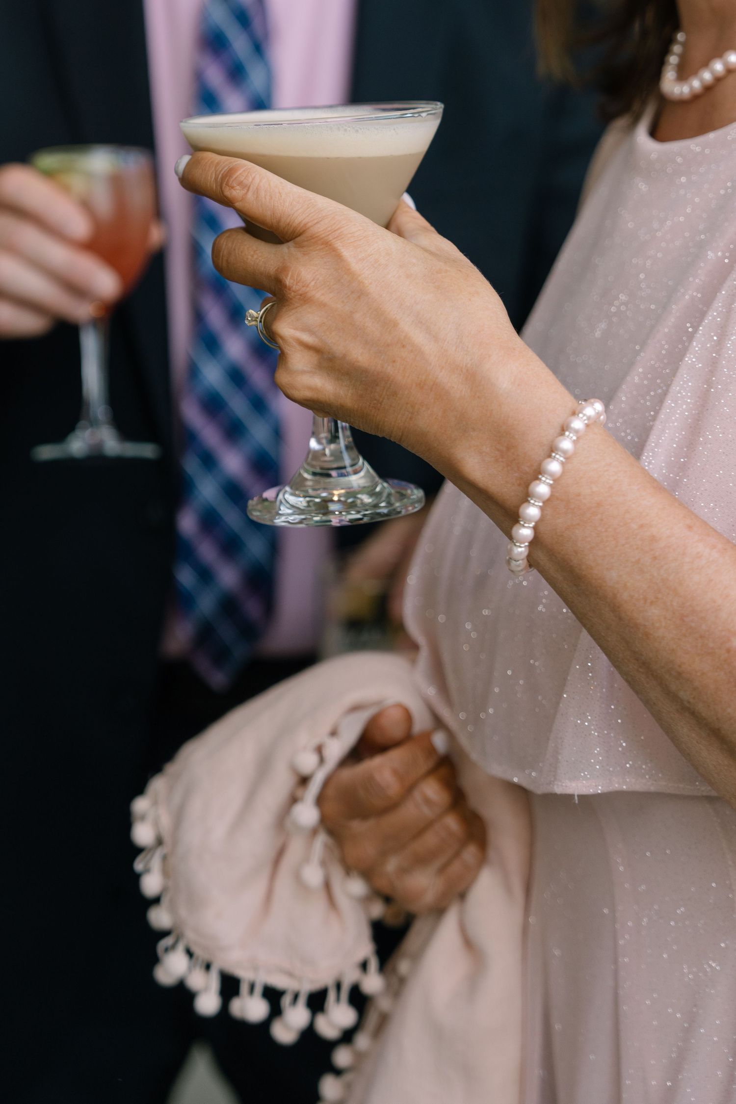 Elegant wedding guest holds a creamy cocktail while wearing a pearl bracelet at a reception.