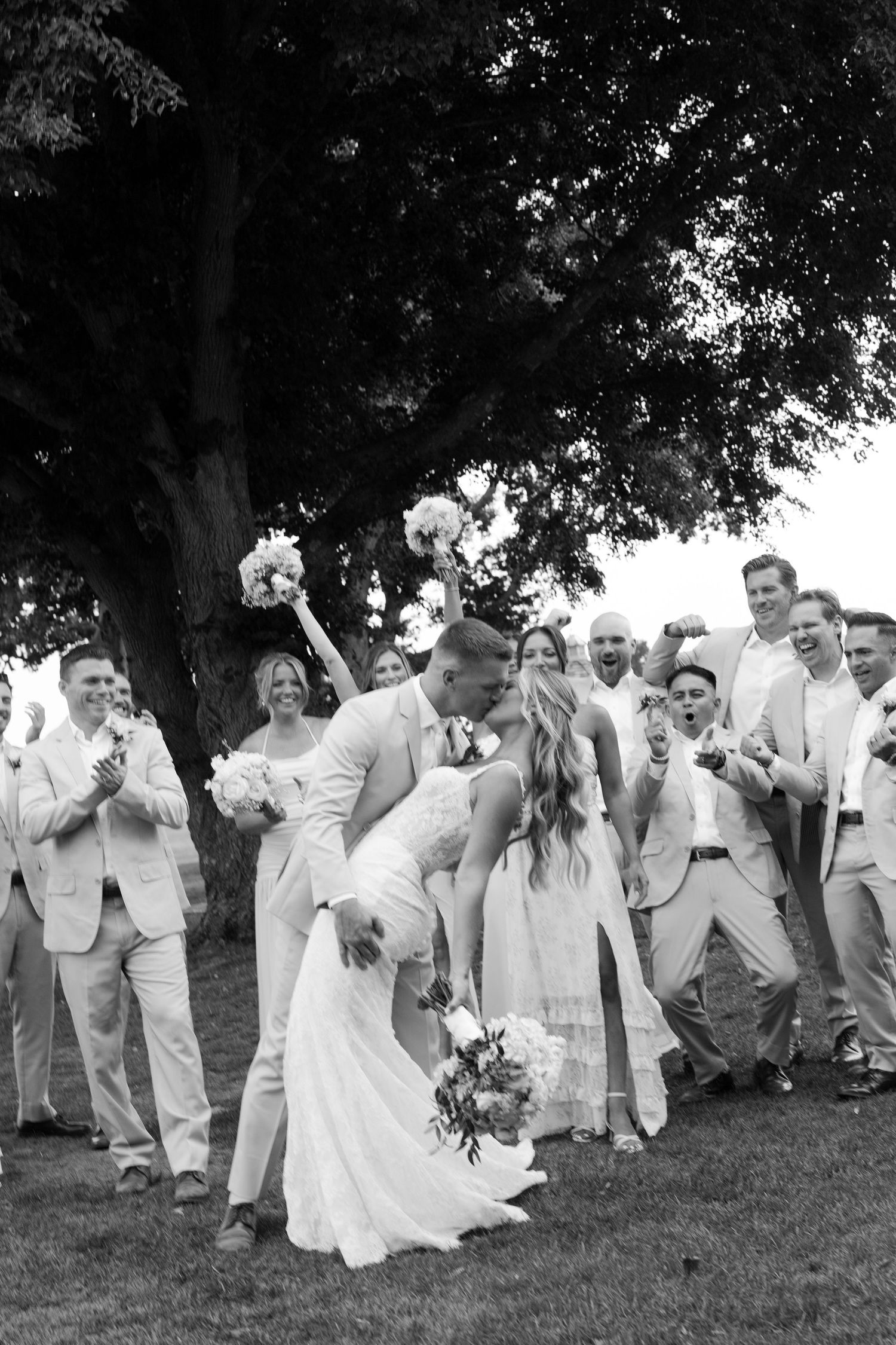 Black and white photo of a groom dipping his bride for a kiss surrounded by cheering wedding party outdoors.