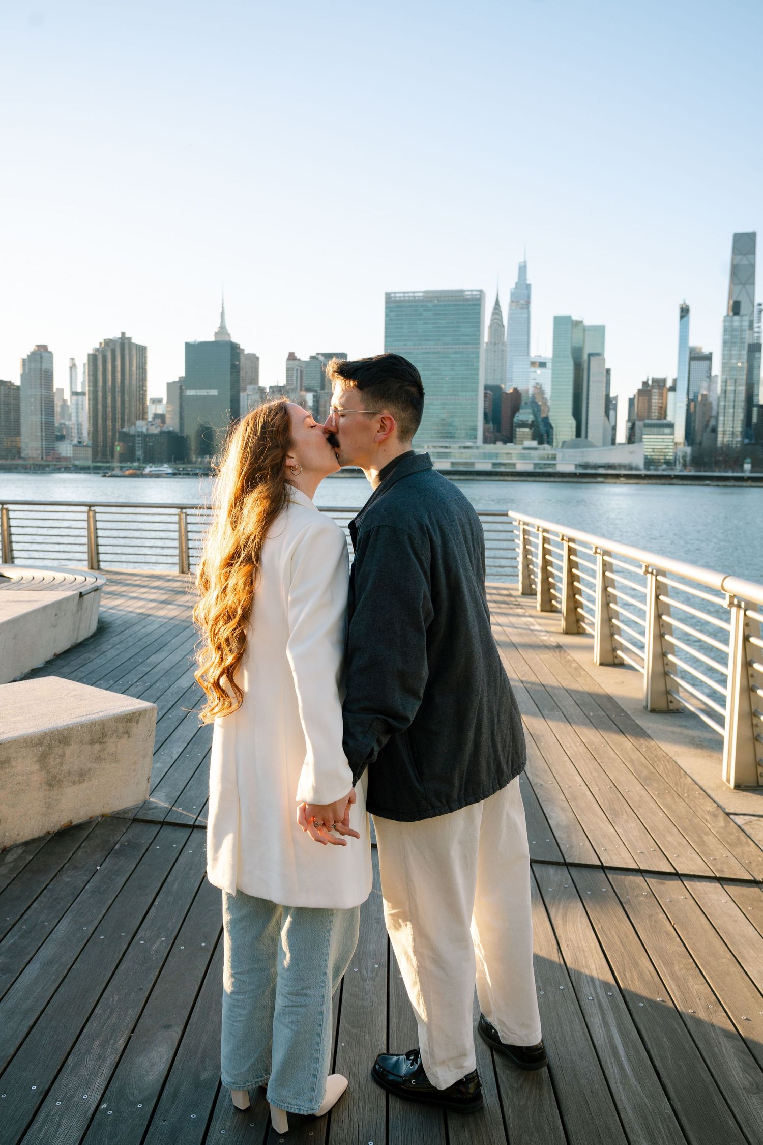 A couple shares a romantic moment on a wooden pier overlooking the Manhattan skyline.