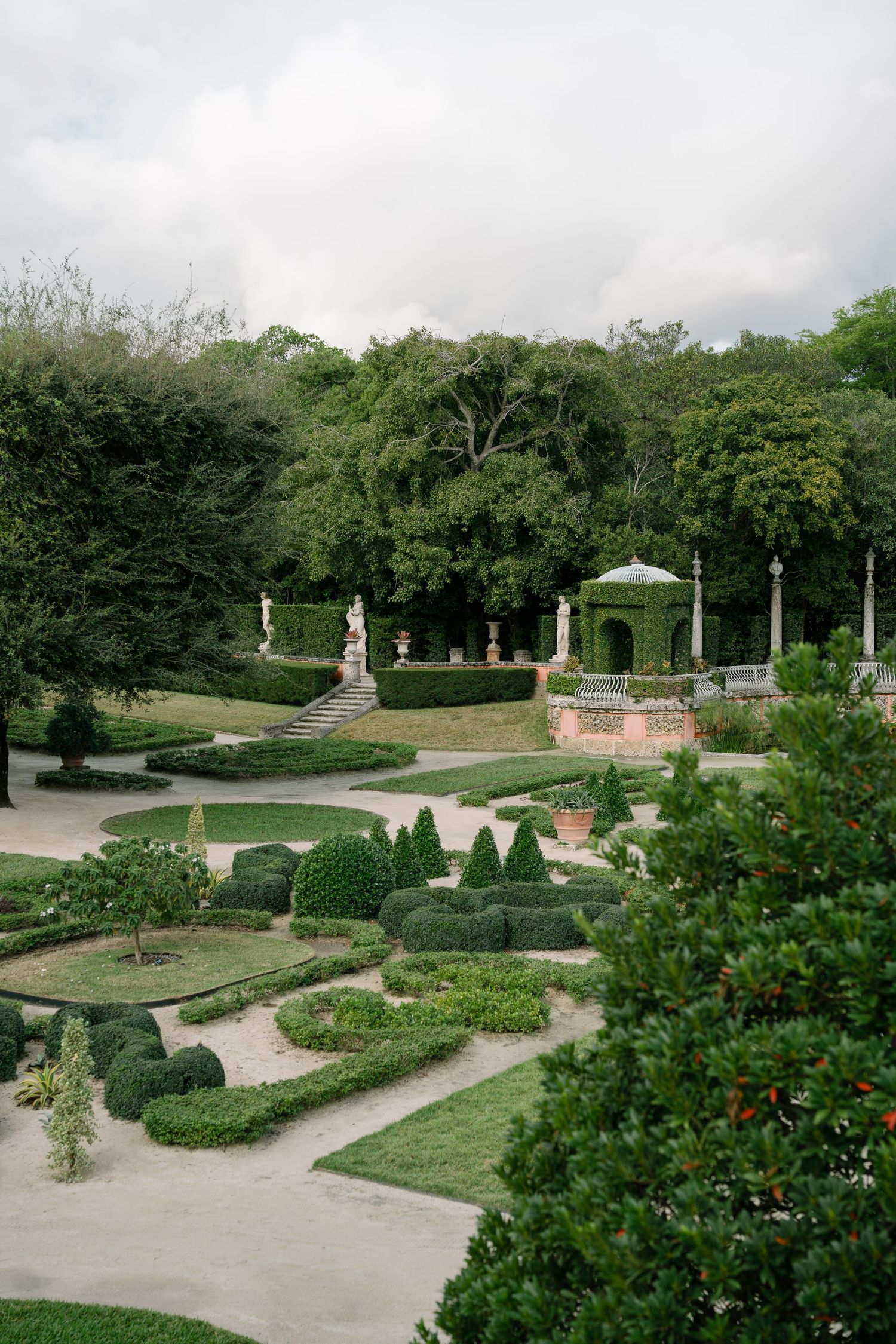 Formal garden with manicured hedges, stone pathways, and a decorative gazebo surrounded by lush greenery.
