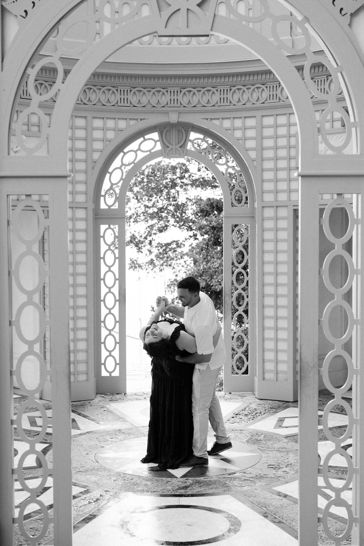 A couple shares an intimate moment under ornate white archways in a classical garden setting.