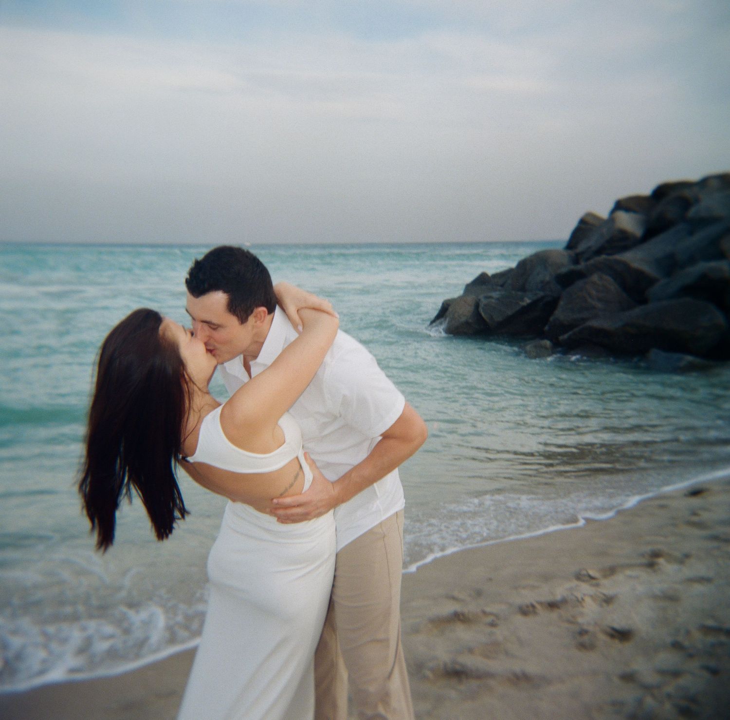 A couple in casual white outfits sharing intimate moments during their beach photoshoot near rocky waters.