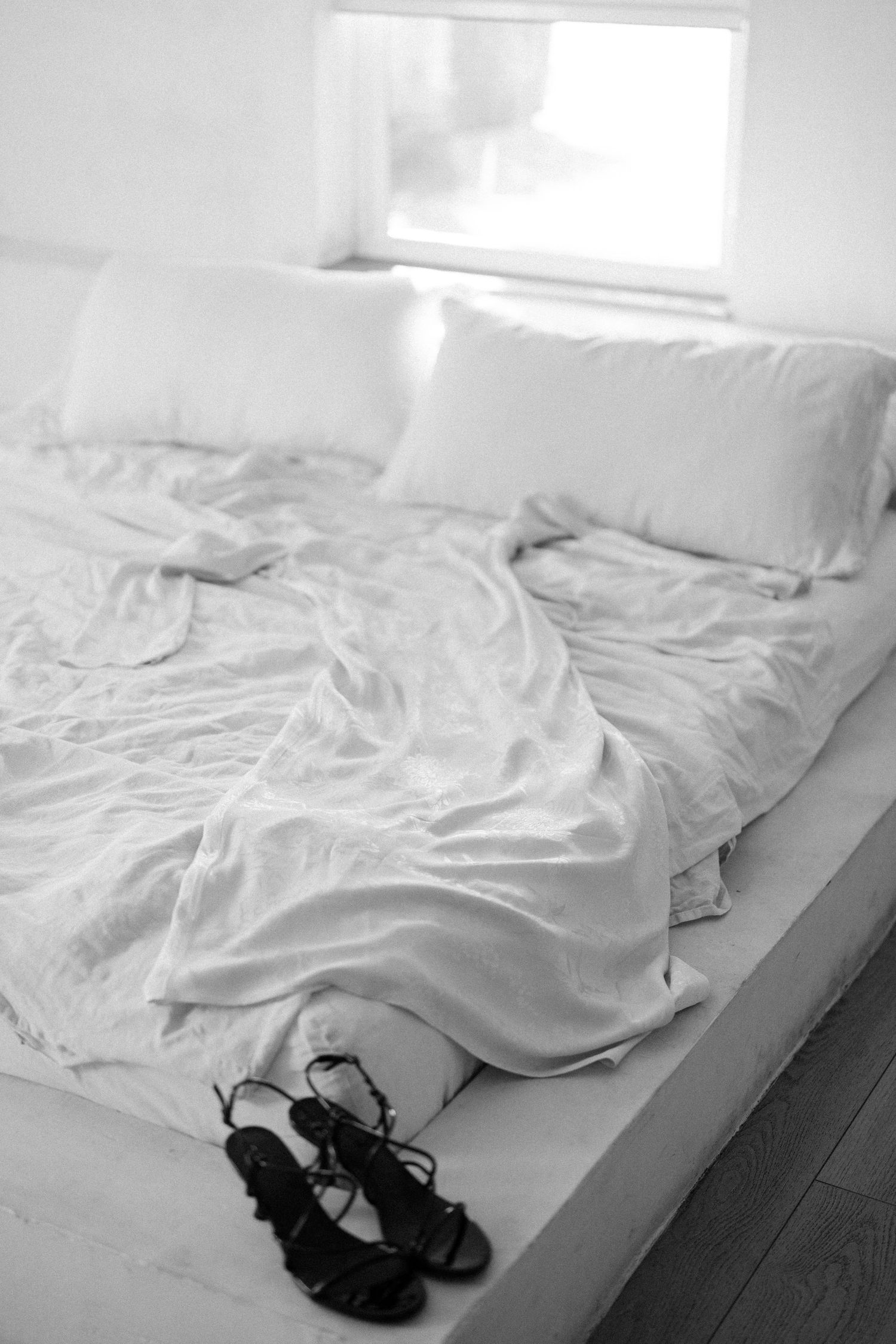 Unmade white bedding with black shoes placed at the end of the bed.