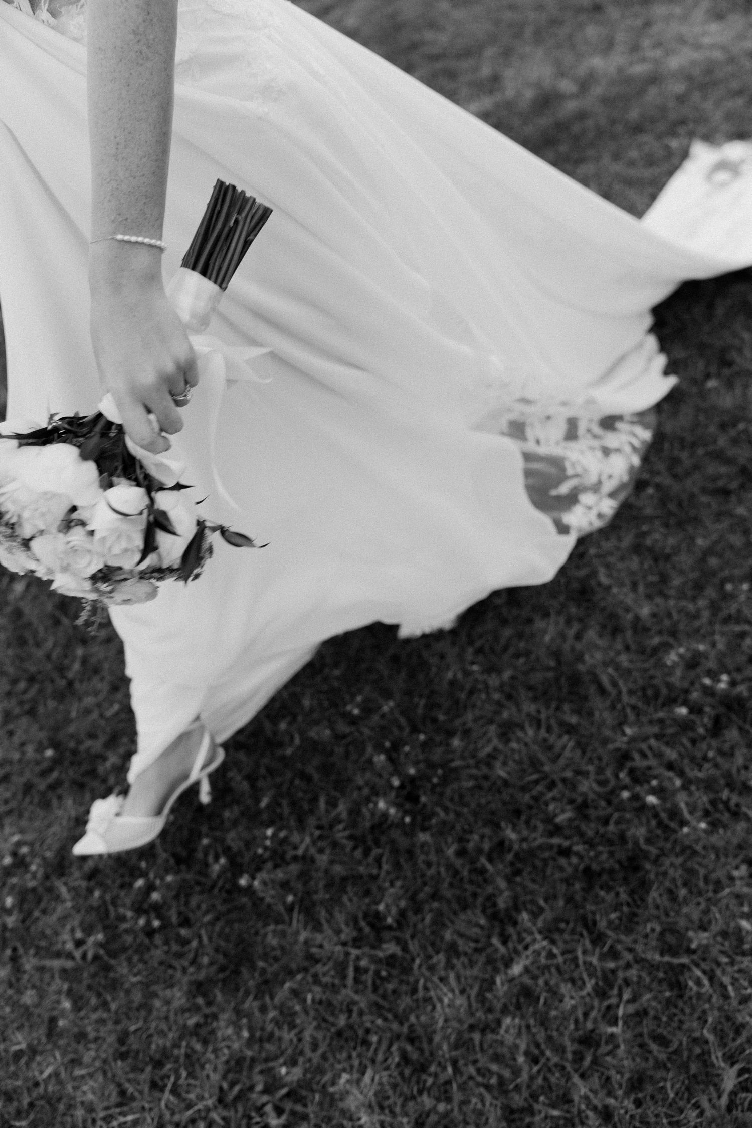 Black and white photo of a flowing white wedding dress hem moving gracefully against grass.