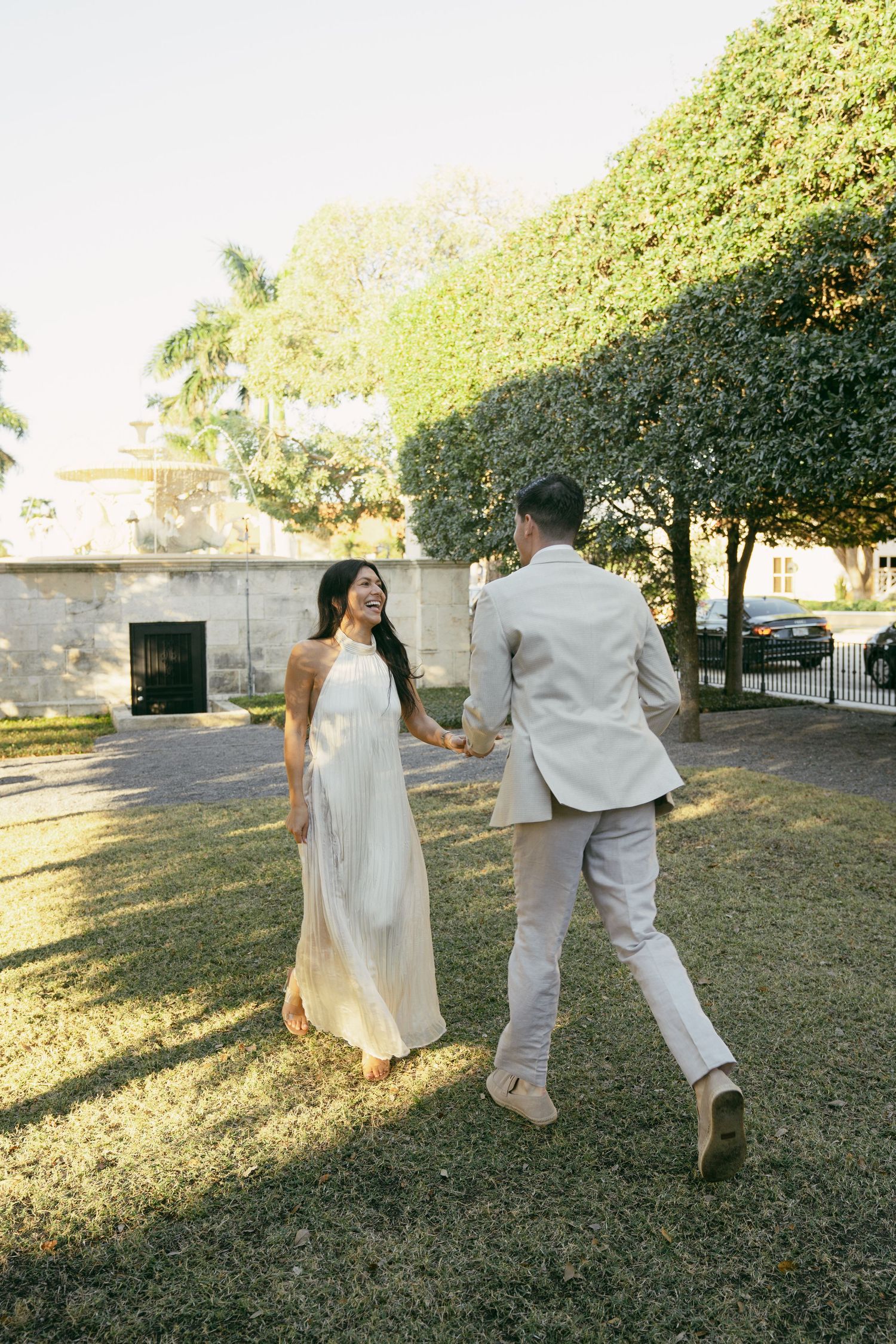 A couple in wedding attire walks together on a grassy lawn surrounded by lush greenery at a historic venue during sunset.