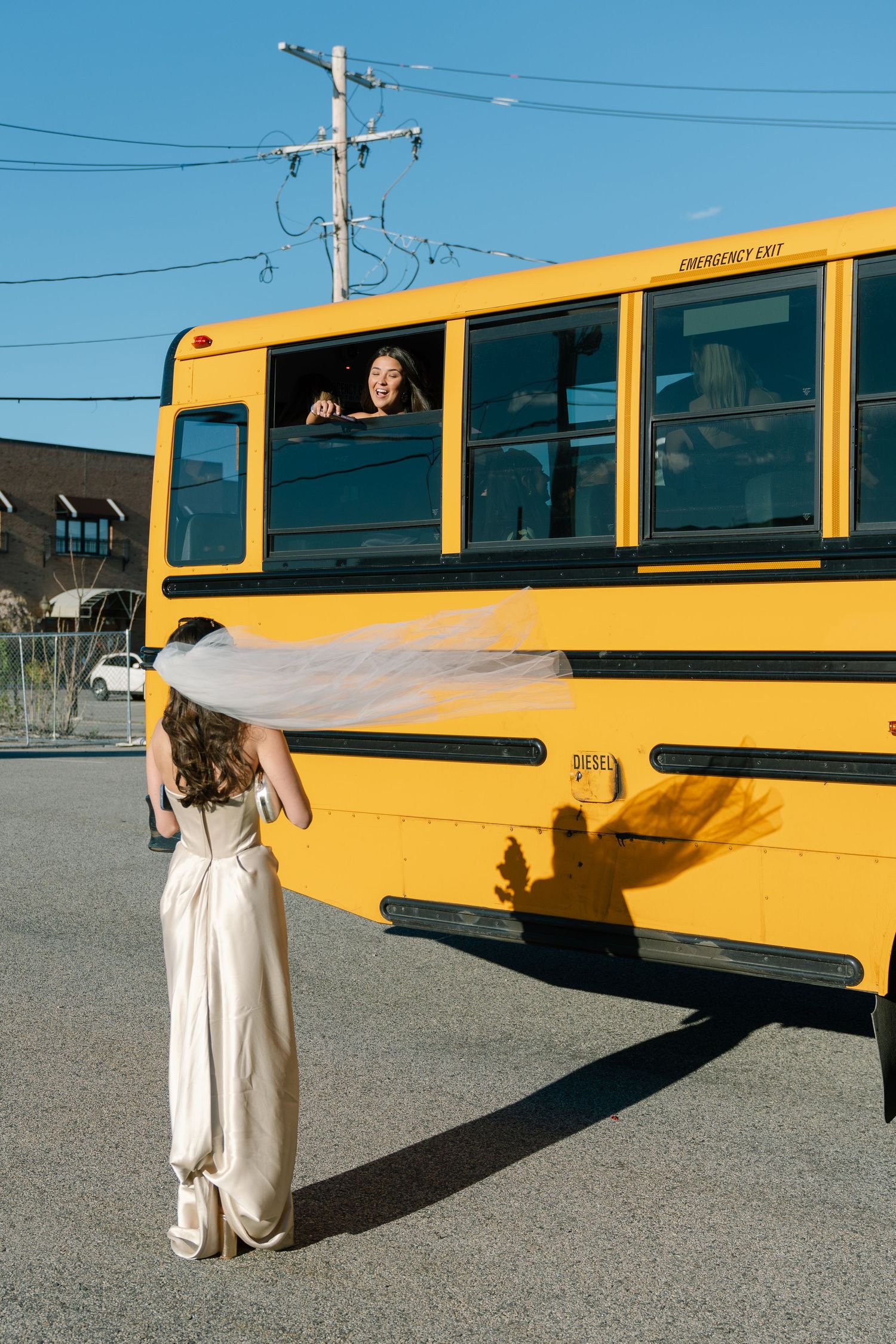 Bride in a white gown with veil blowing in the wind standing beside a yellow school bus outdoors.