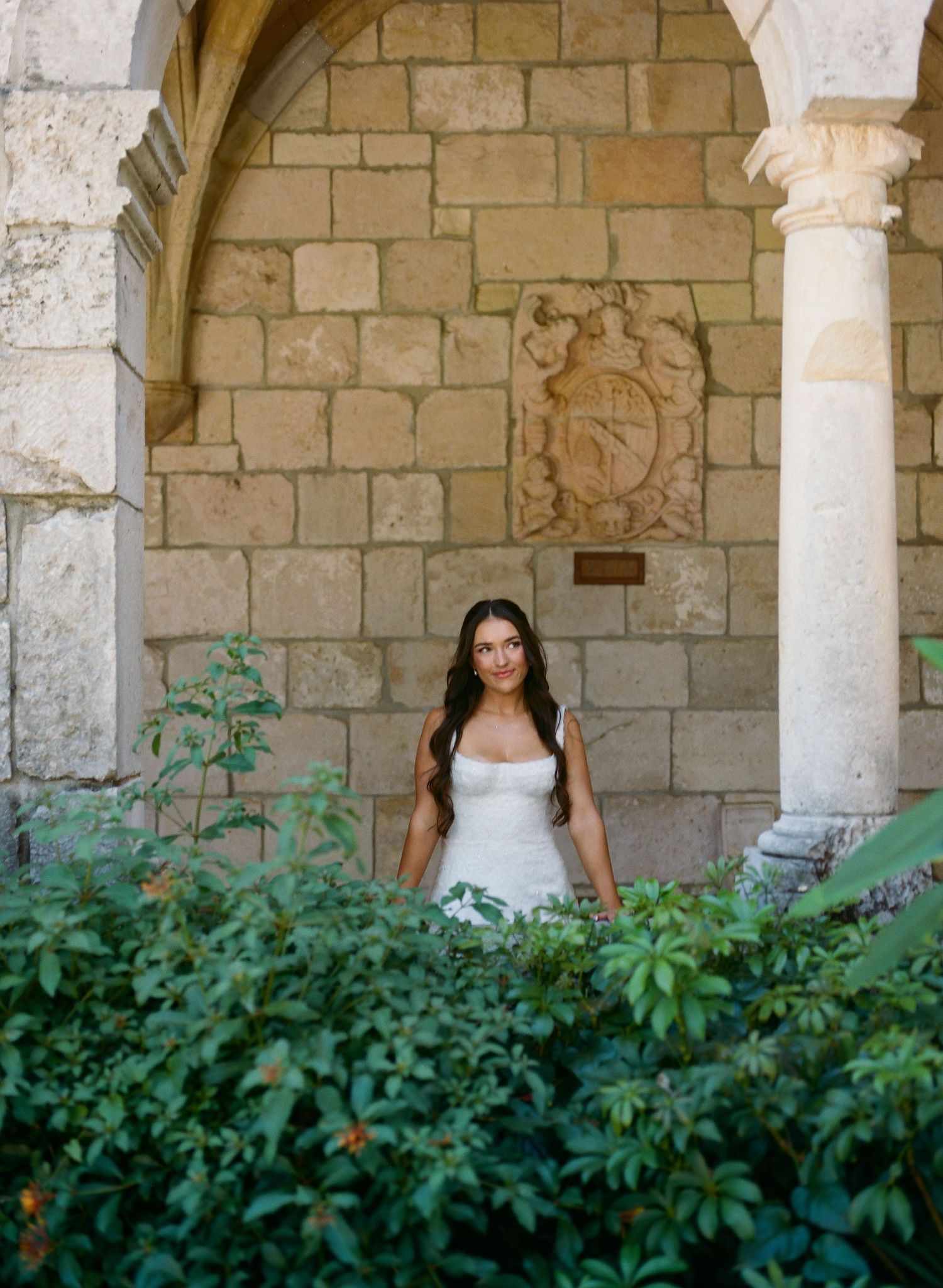 Bride in sleek white gown poses under a stone archway surrounded by lush green foliage.