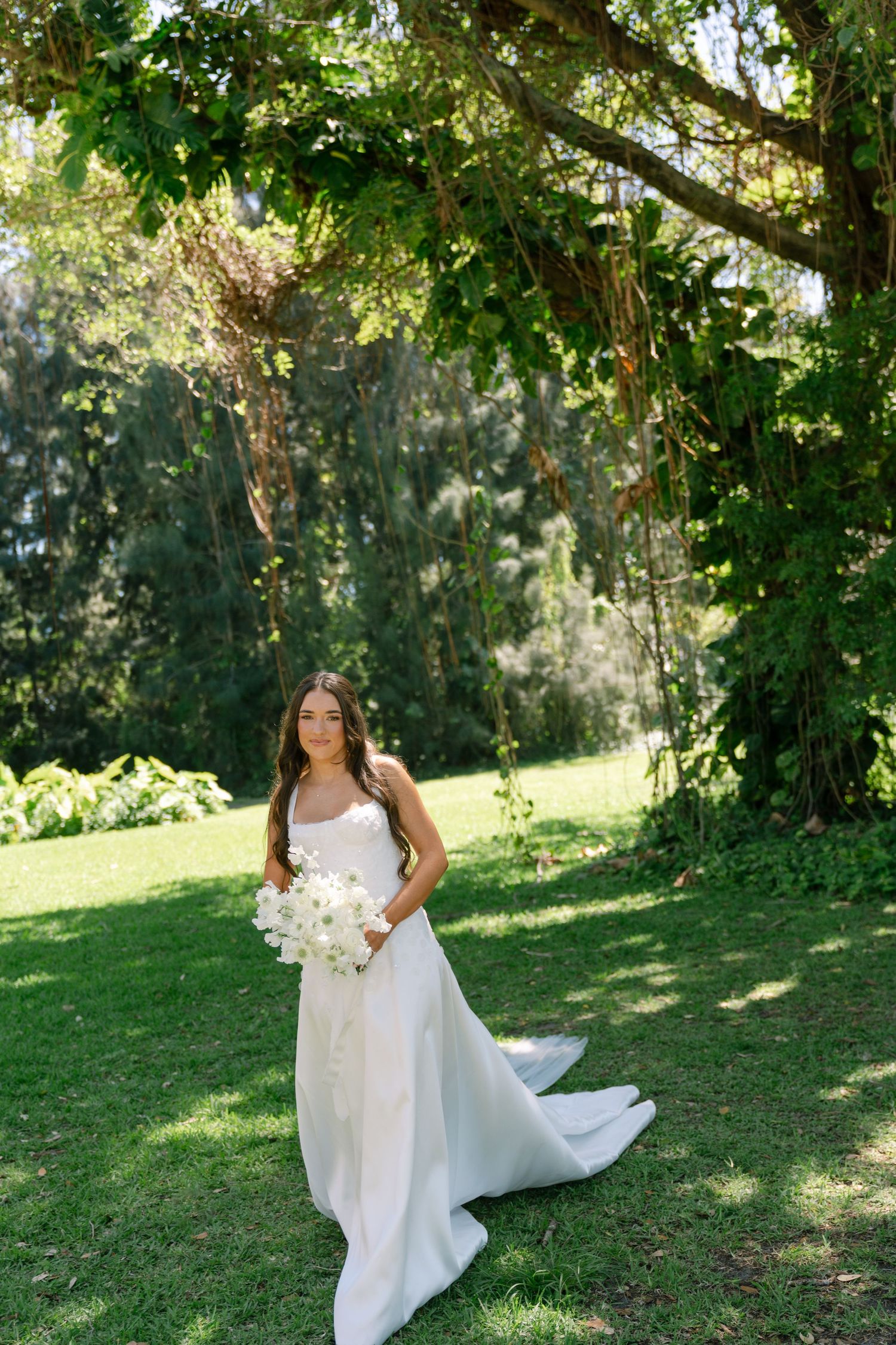 Bride in elegant white gown holding white bouquet standing on green lawn under lush tree canopy.