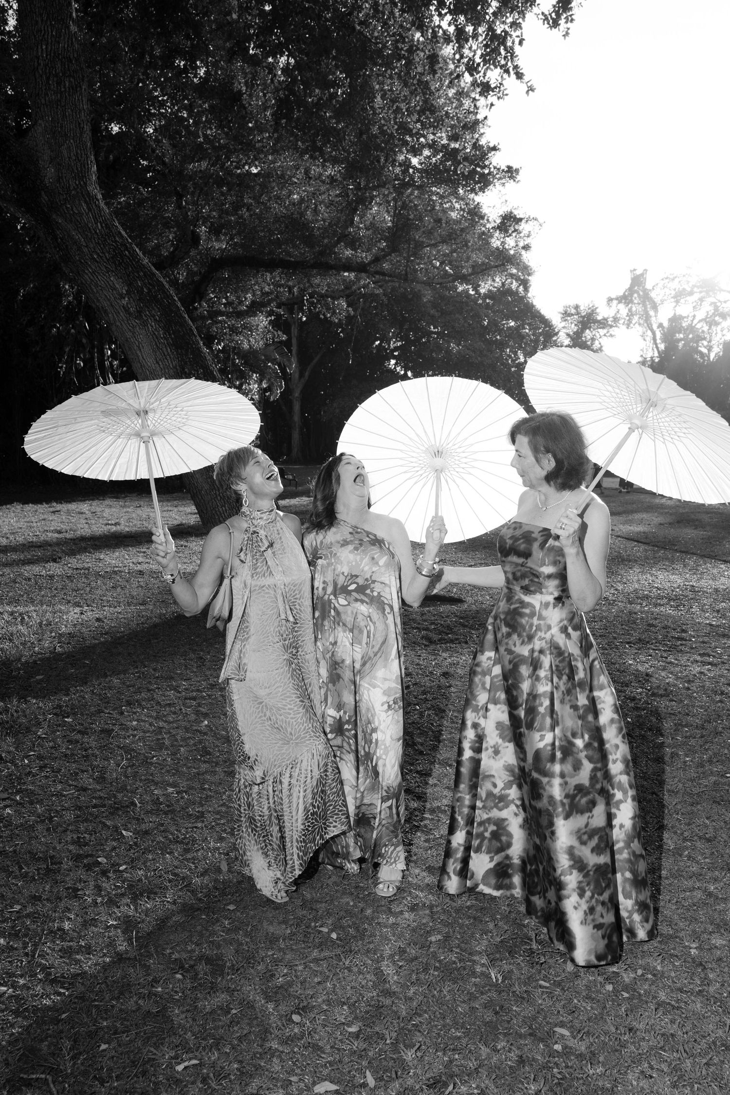 Three women in elegant gowns holding parasols laughing outdoors in black and white photo.