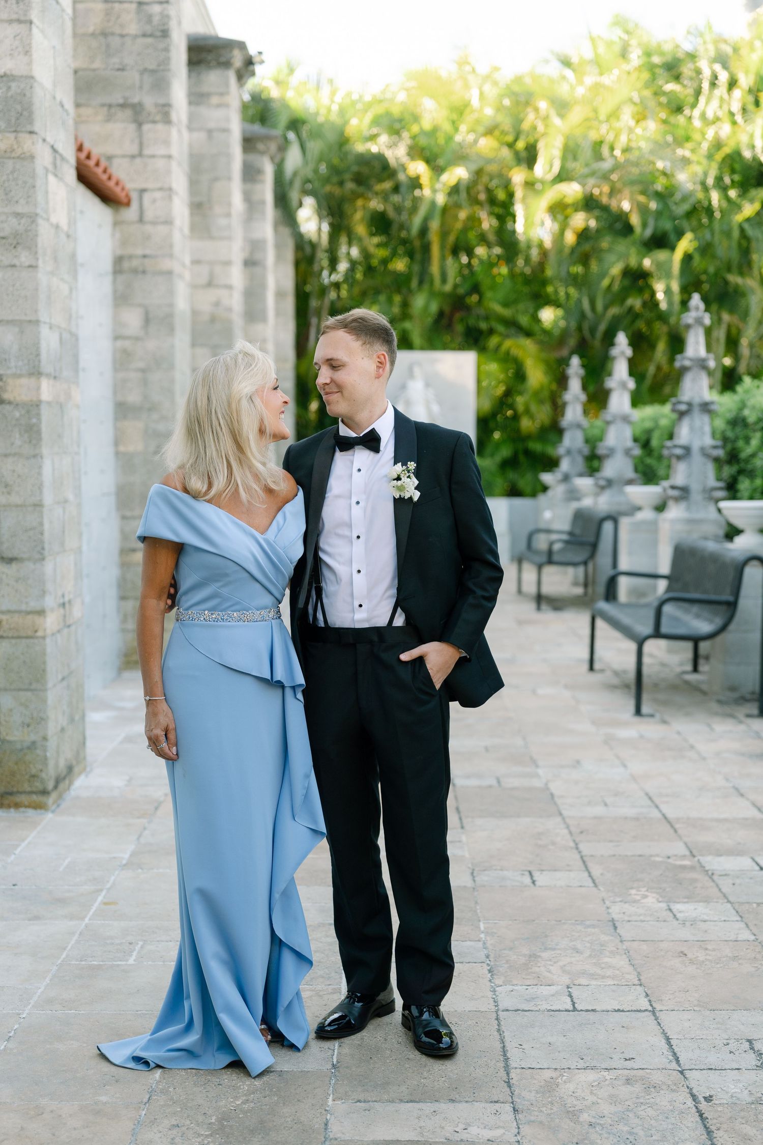 Groom in black tuxedo with mother in blue off-shoulder gown posing at stone wedding venue.