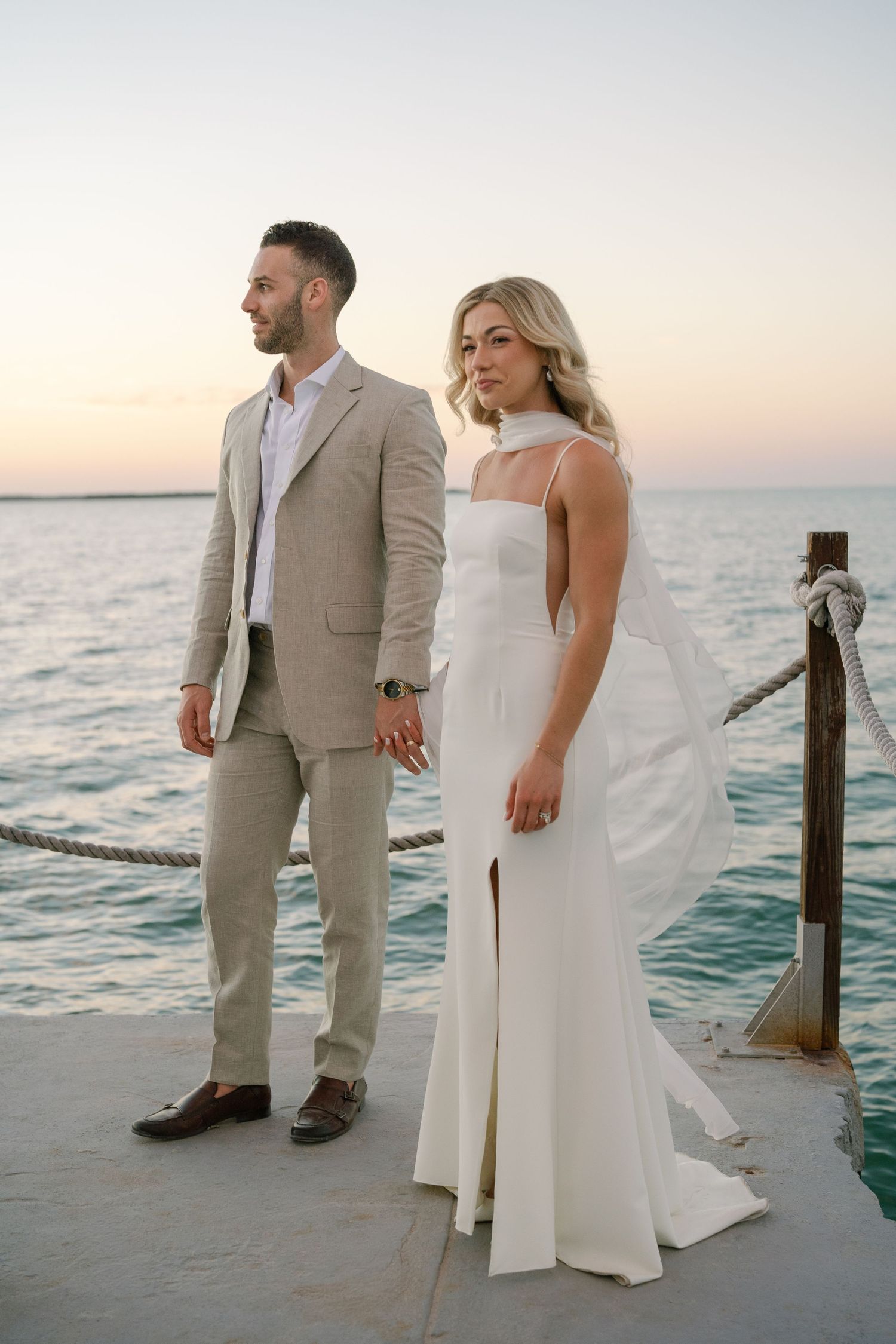 A couple in wedding attire stands together on a dock overlooking the ocean at dusk.