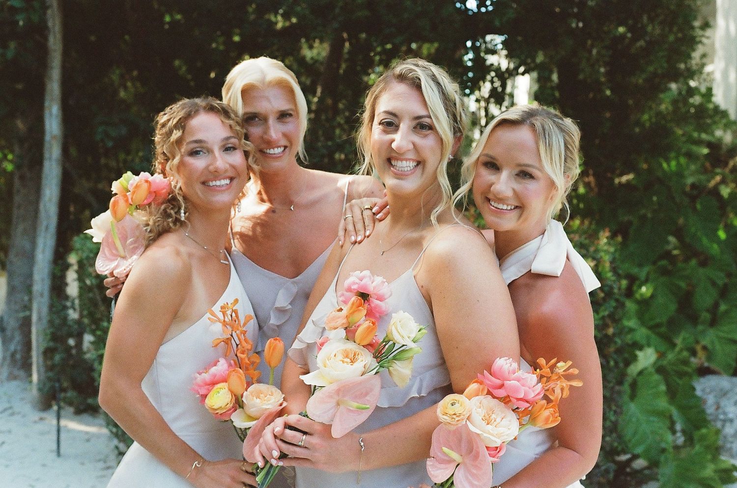 Four bridesmaids in gray dresses hold peach and coral colored bouquets while smiling outdoors at a wedding.