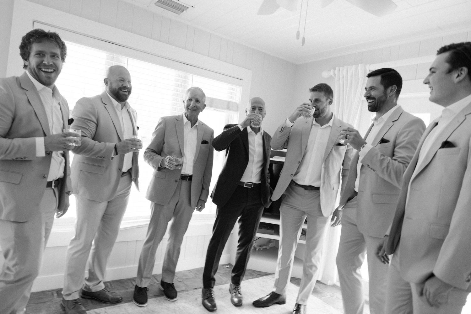 Groomsmen in light colored suits gather in a line during wedding preparations.