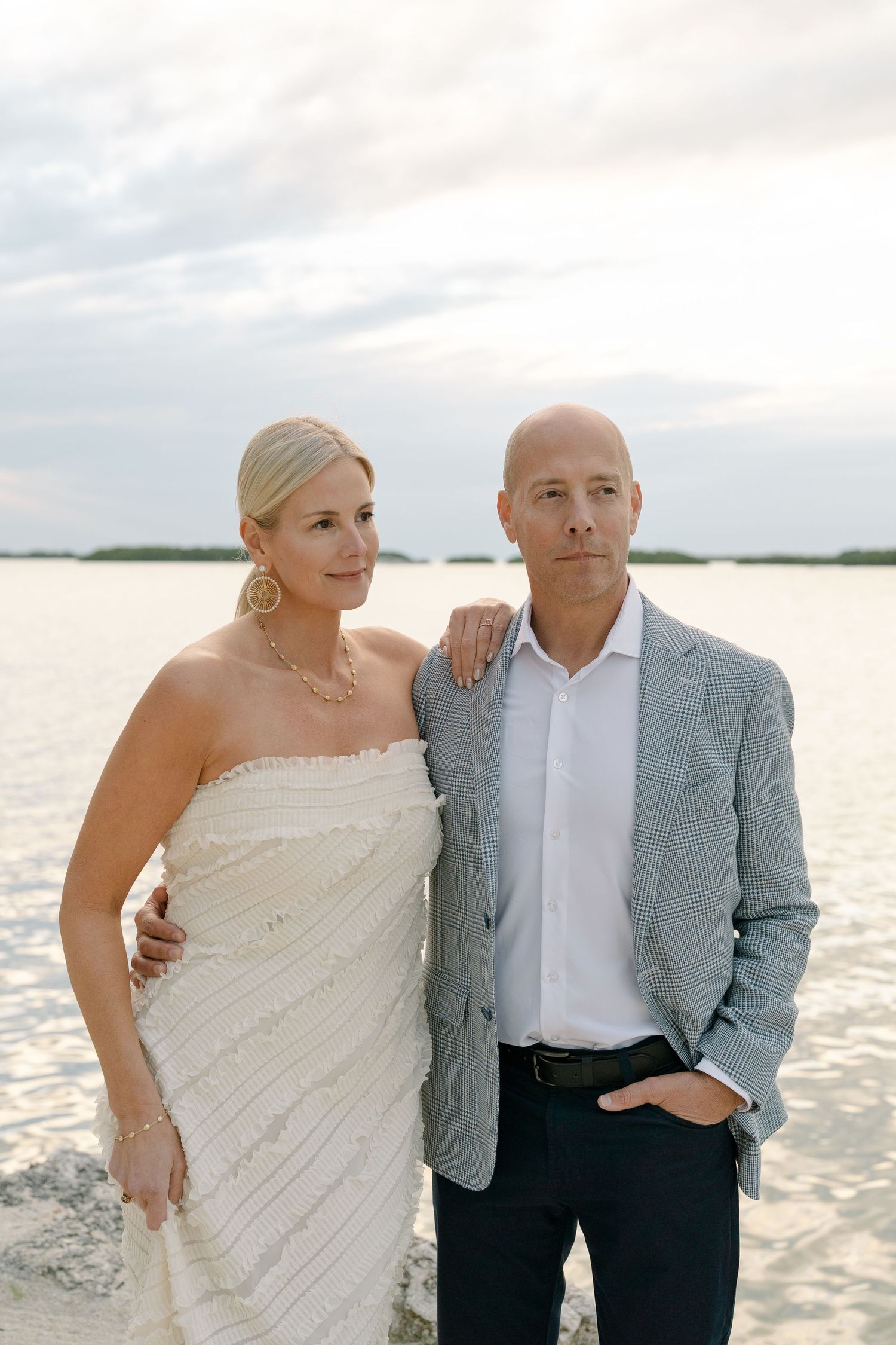A couple poses for romantic beach wedding photos at sunset in elegant white and gray attire.
