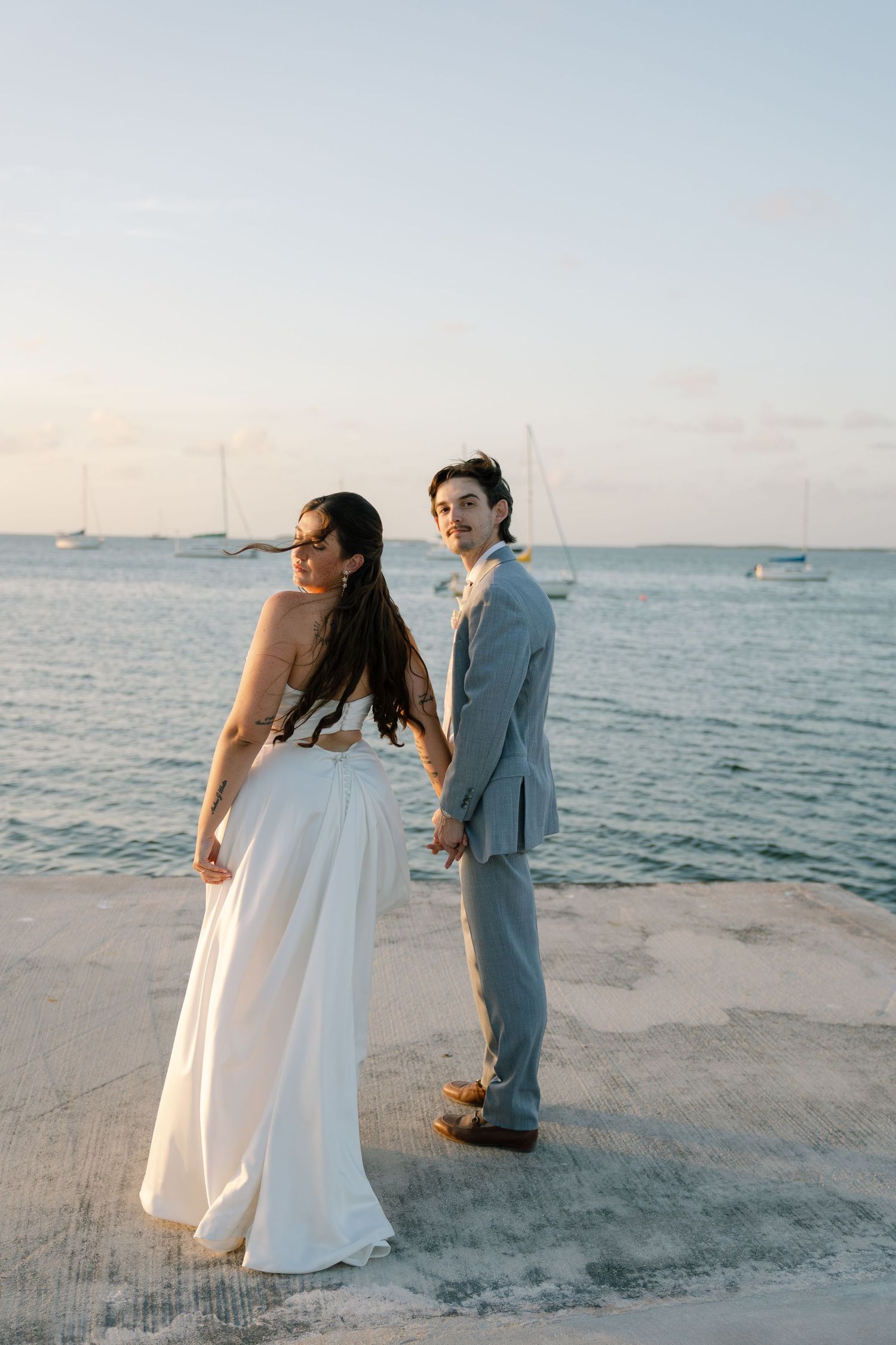A couple in wedding attire shares a romantic moment on a sandy beach with sailboats visible in the ocean background.