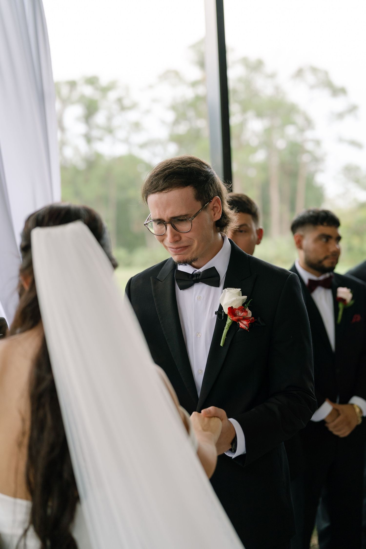 Emotional groom in black tuxedo with boutonniere holds bride's hands during outdoor wedding ceremony.