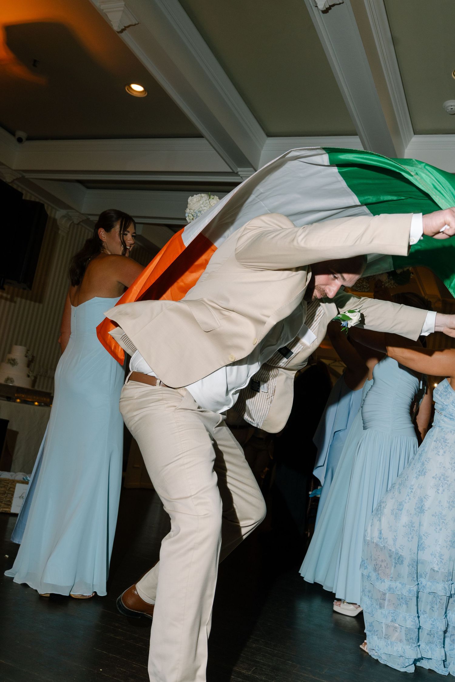 Groom in beige suit dancing energetically with Irish flag at lively wedding reception.