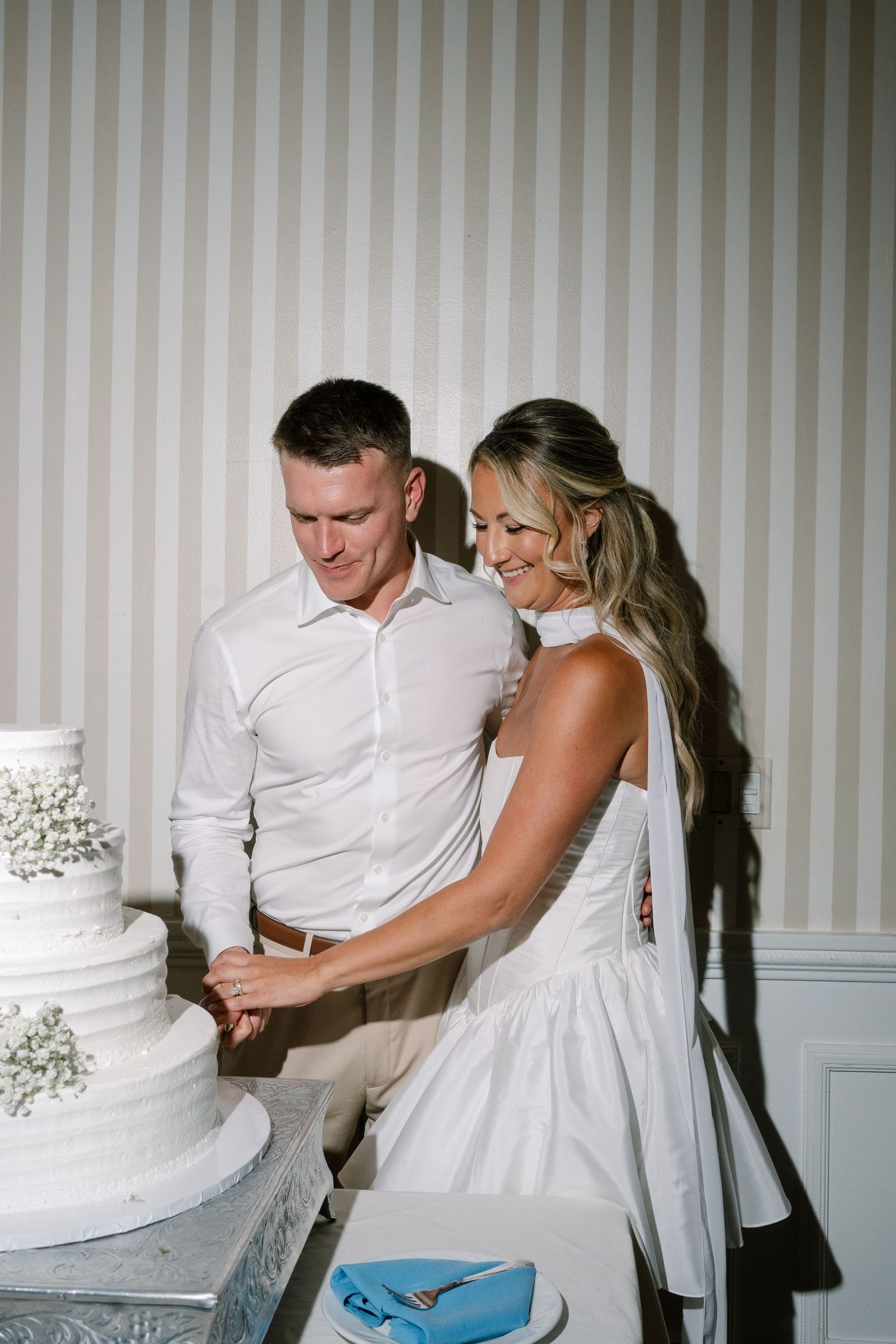 Bride and groom cutting white tiered wedding cake together indoors against striped wallpaper background.