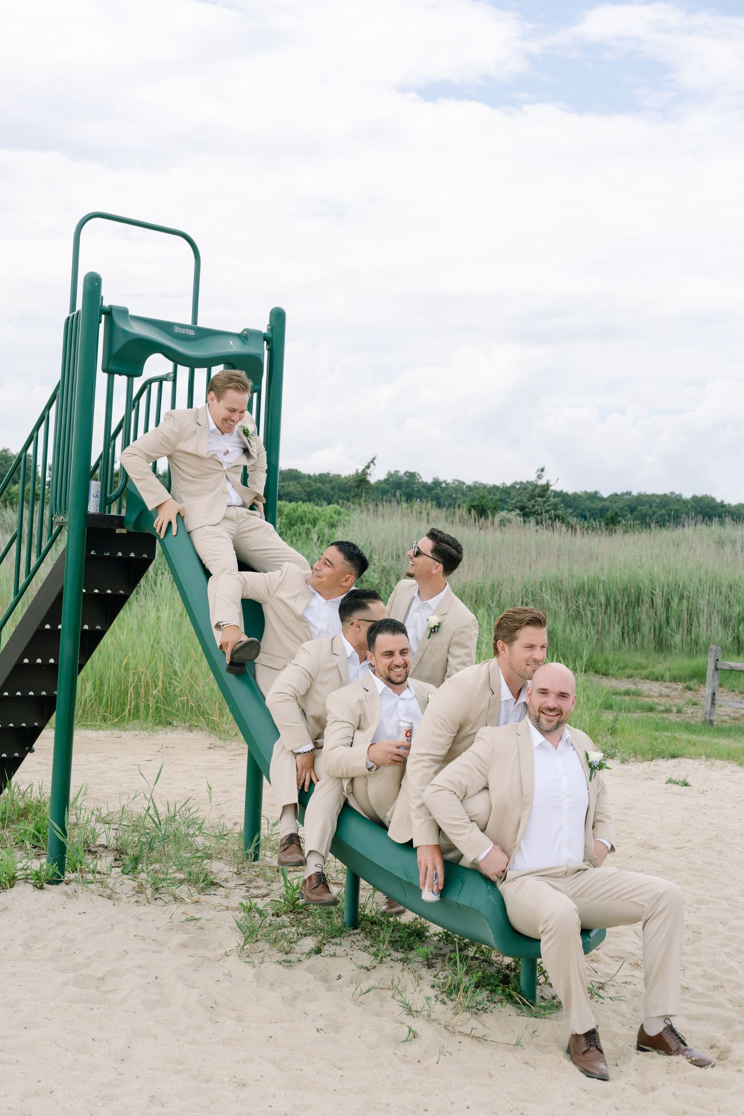 Groomsmen in tan suits posing playfully on a green playground slide at a beach wedding venue.