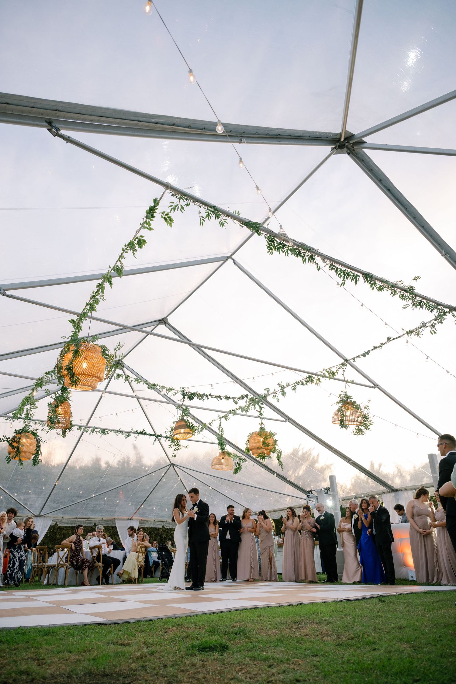 Couples share first dance under a clear tent adorned with hanging lanterns and greenery at a wedding.
