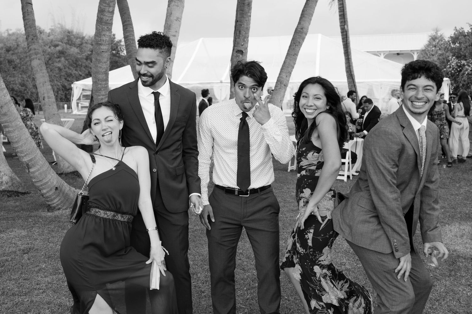Black and white photo of five wedding guests smiling and posing playfully between palm trees.