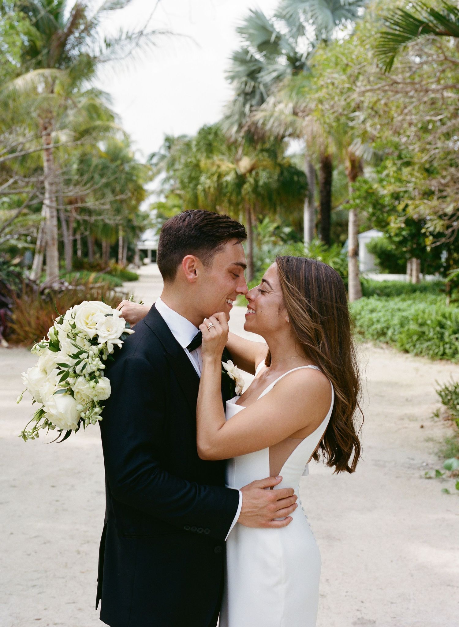 Bride and groom share a romantic moment surrounded by palm trees holding white floral bouquet.
